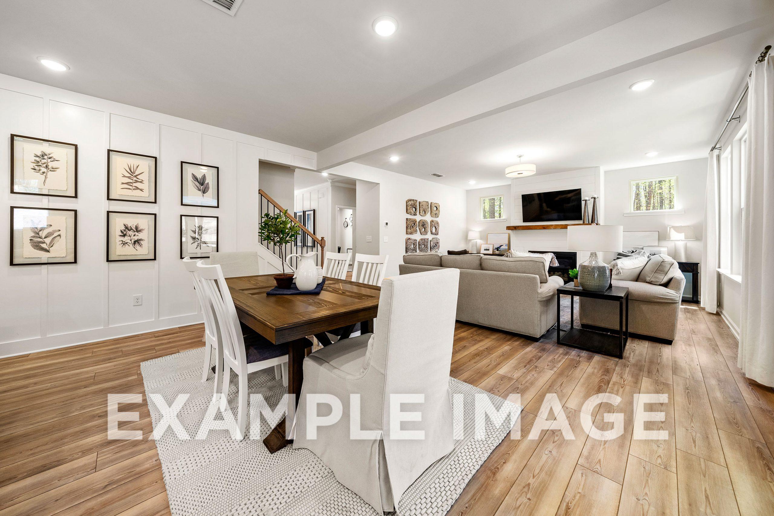 Open-concept living and dining in The Hickory B with wooden table, white chairs, L-shaped sofa, fireplace, TV, and staircase
