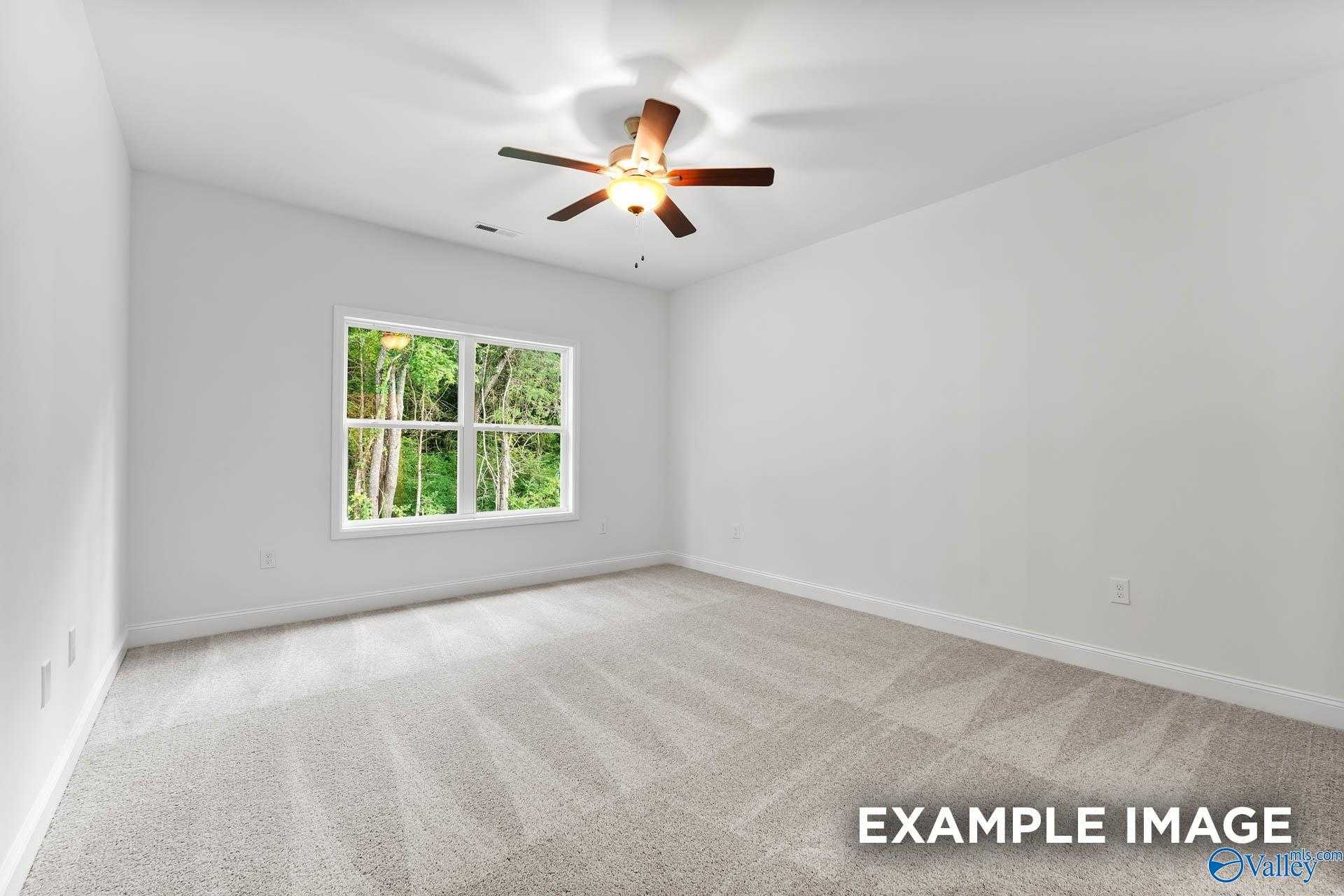 Bright empty bedroom with white walls, beige carpet, ceiling fan, and large windows showing greenery in Davidson Homes The Daphne V, Athens, Alabama