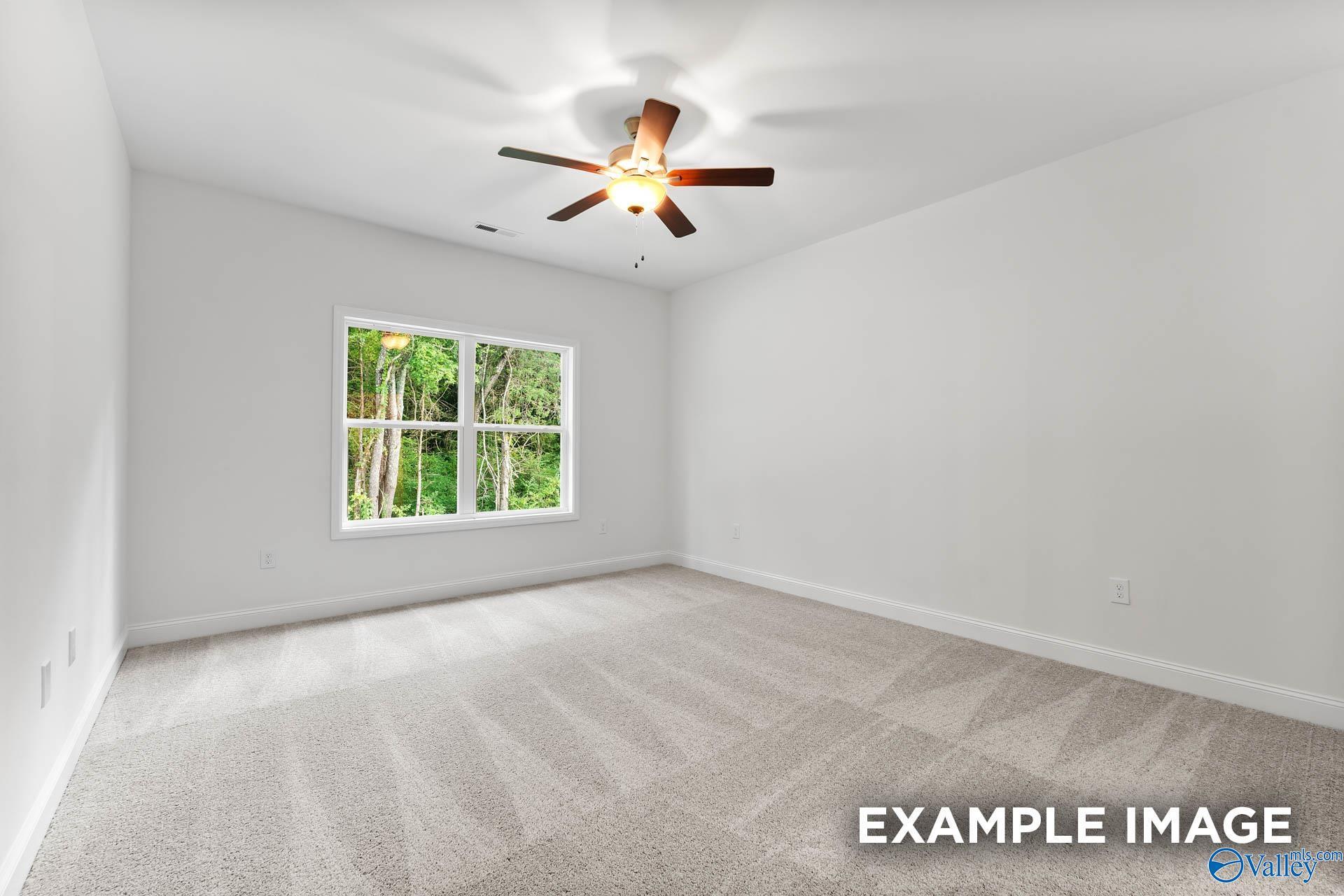 Bright empty bedroom with white walls, beige carpet, ceiling fan, and large windows showing greenery in Davidson Homes The Daphne V, Athens, Alabama