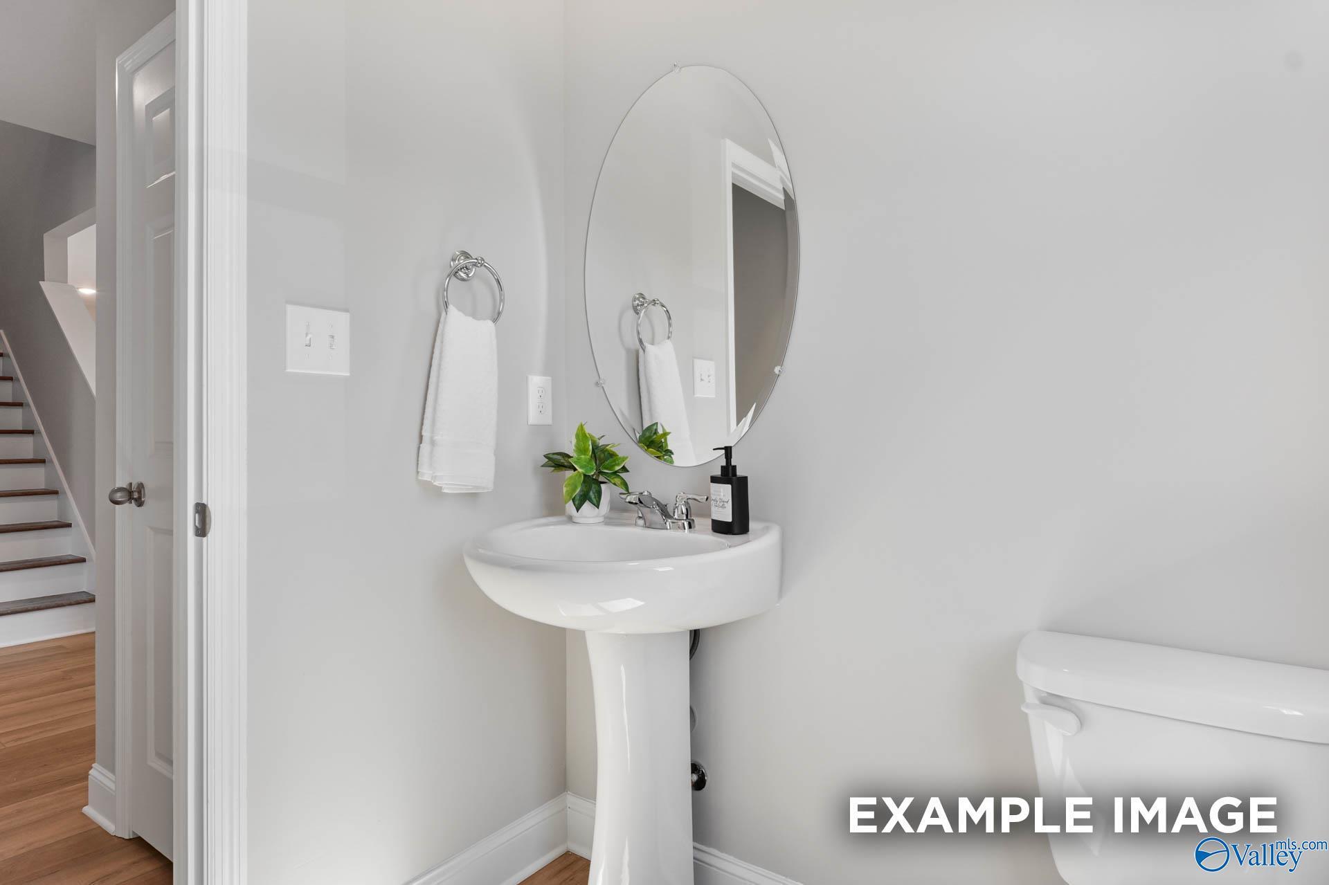 Elegant powder room featuring white pedestal sink, round mirror, and toilet in Davidson Homes The Chelsea B, Toney, Alabama