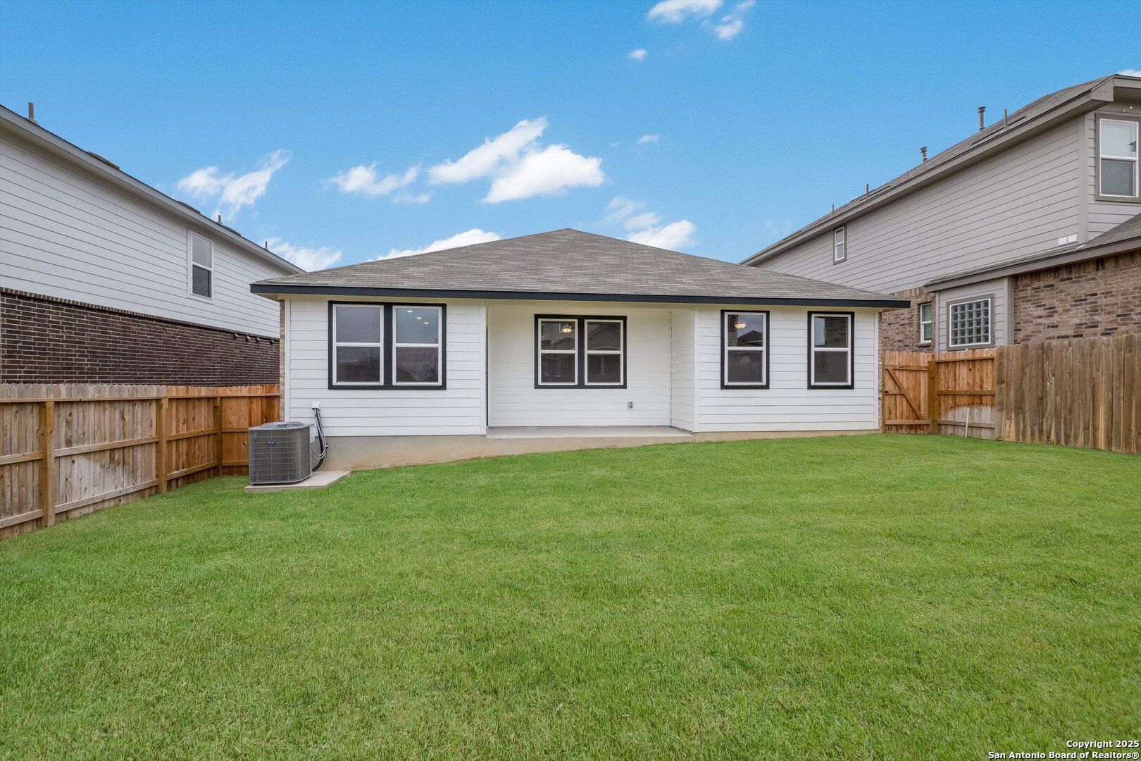 Rear view of single-story Asheville K home with white siding, black windows, covered patio, lush green yard, and wooden fence in Bricewood, San Antonio