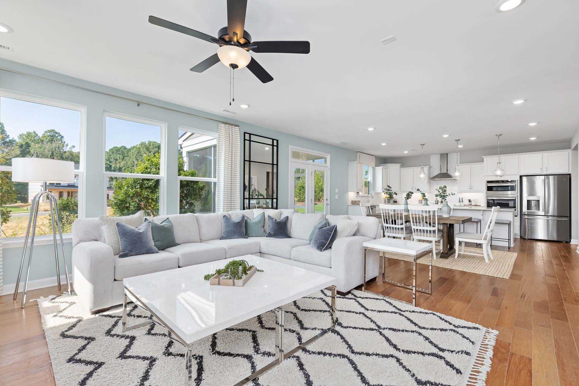 Spacious open-concept living room at Bentwinds in Fuquay-Varina NC with hardwood floors, gray sectional sofa, ceiling fans, and white kitchen by Davidson Homes