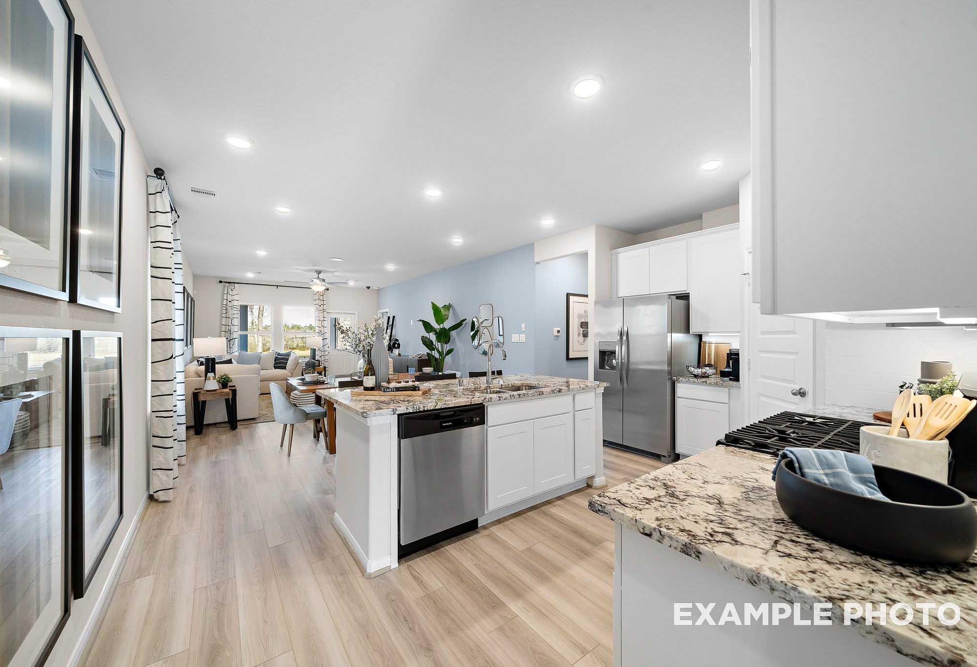 Open-concept kitchen in The San Marcos home design with white cabinets, granite island, stainless appliances, and adjacent living space
