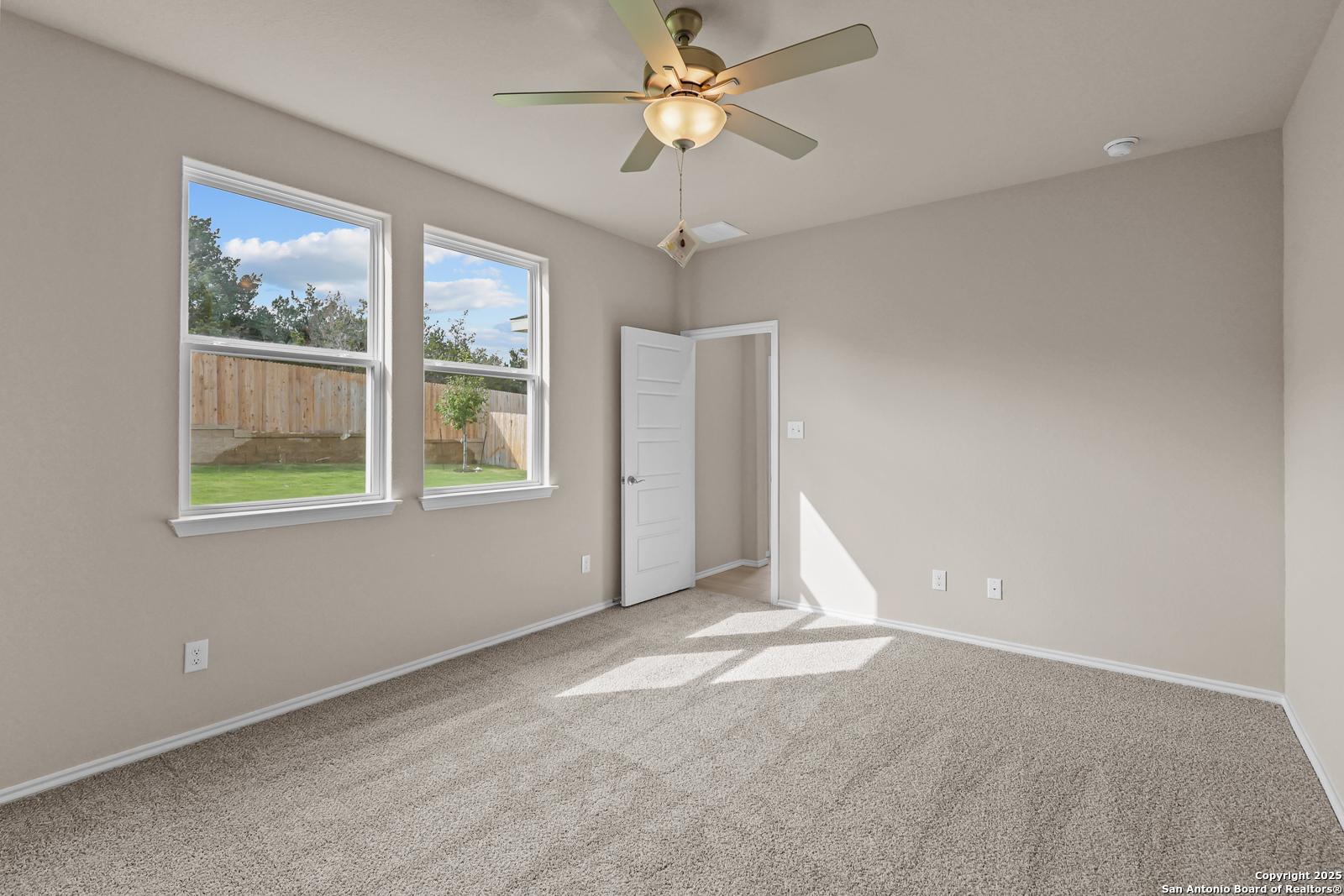 Bright bedroom with beige walls, large windows to lush backyard, ceiling fan and white door in Davidson Homes The Douglas G, San Antonio