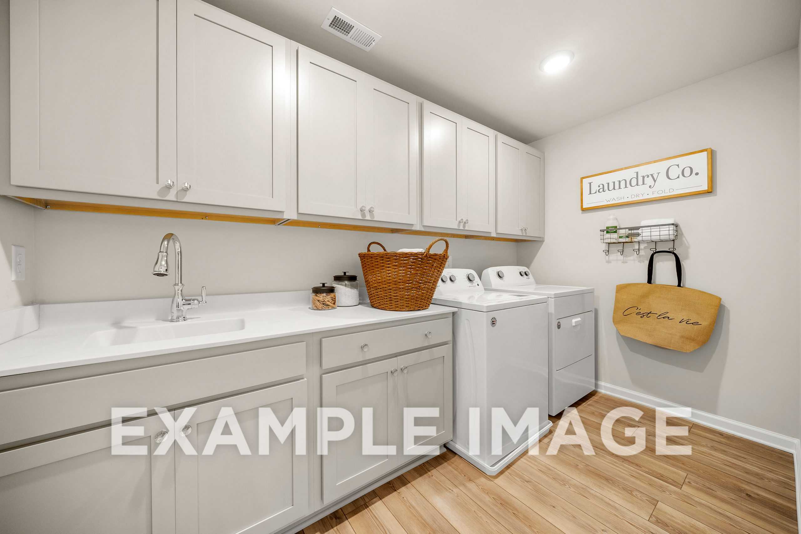 Spacious laundry room in The Hickory A home with white cabinets, utility sink, washer dryer, and wicker storage