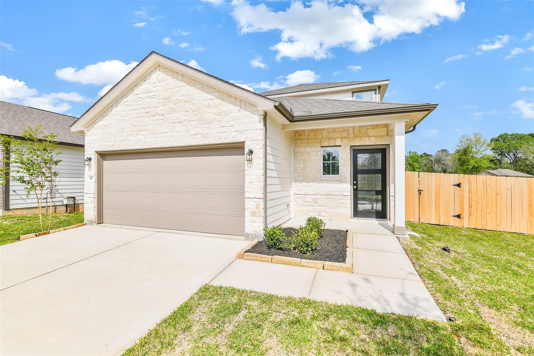Modern 2-story San Marcos E home exterior with beige stone facade, 2-car garage, glass door, and landscaped yard in Conroe, Texas