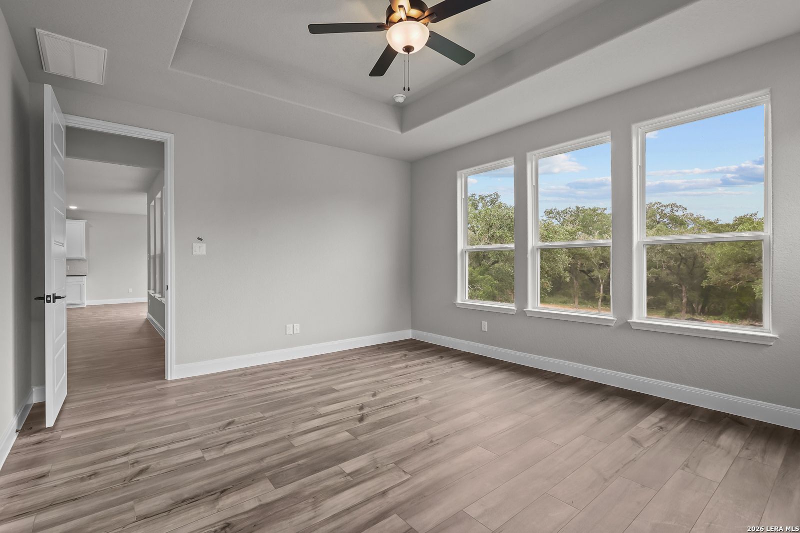 Bright bedroom with light gray walls, hardwood floors, ceiling fan, and large windows overlooking trees in Davidson Homes The Belmont H, Castroville, Texas