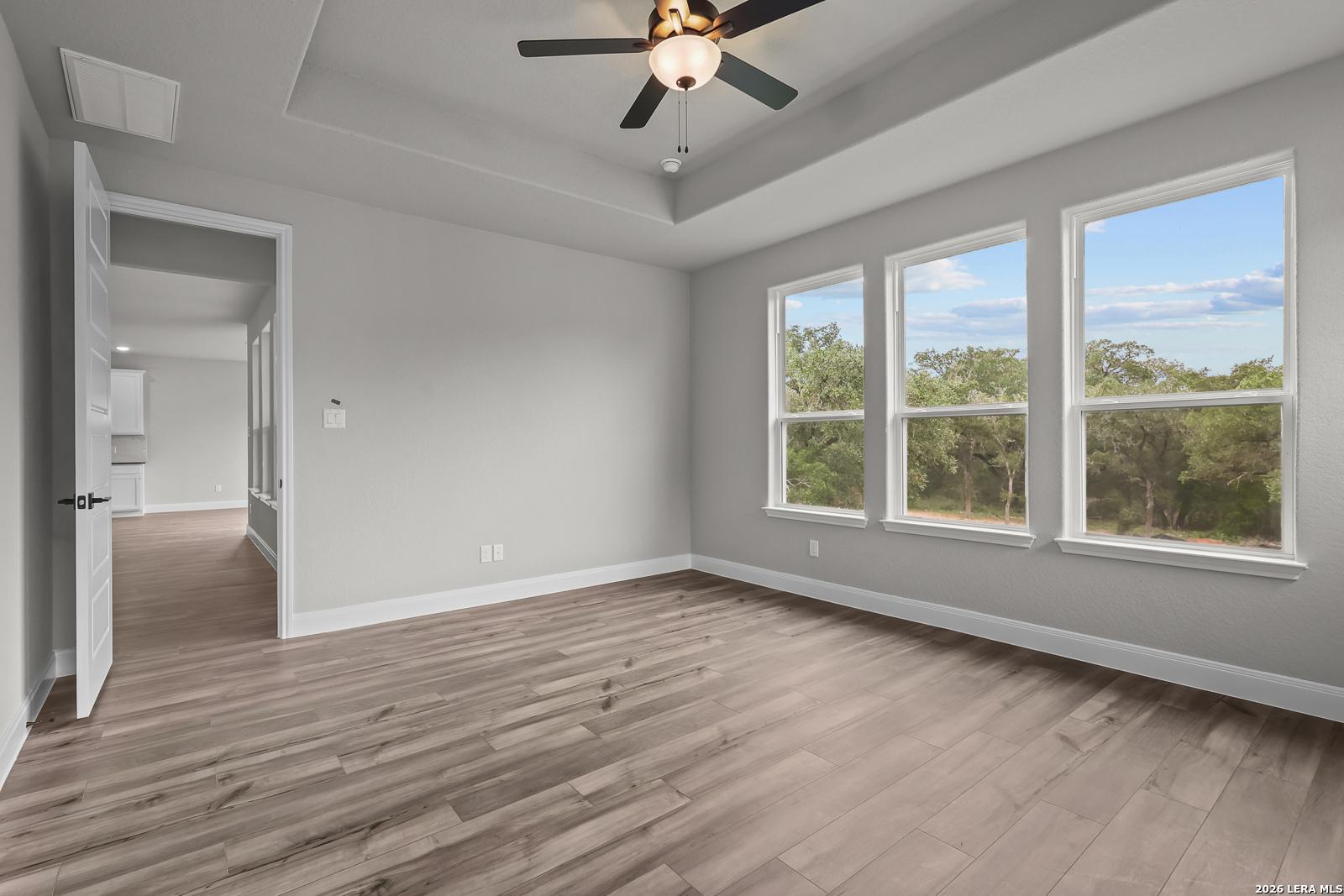 Bright bedroom with light gray walls, hardwood floors, ceiling fan, and large windows overlooking trees in Davidson Homes The Belmont H, Castroville, Texas