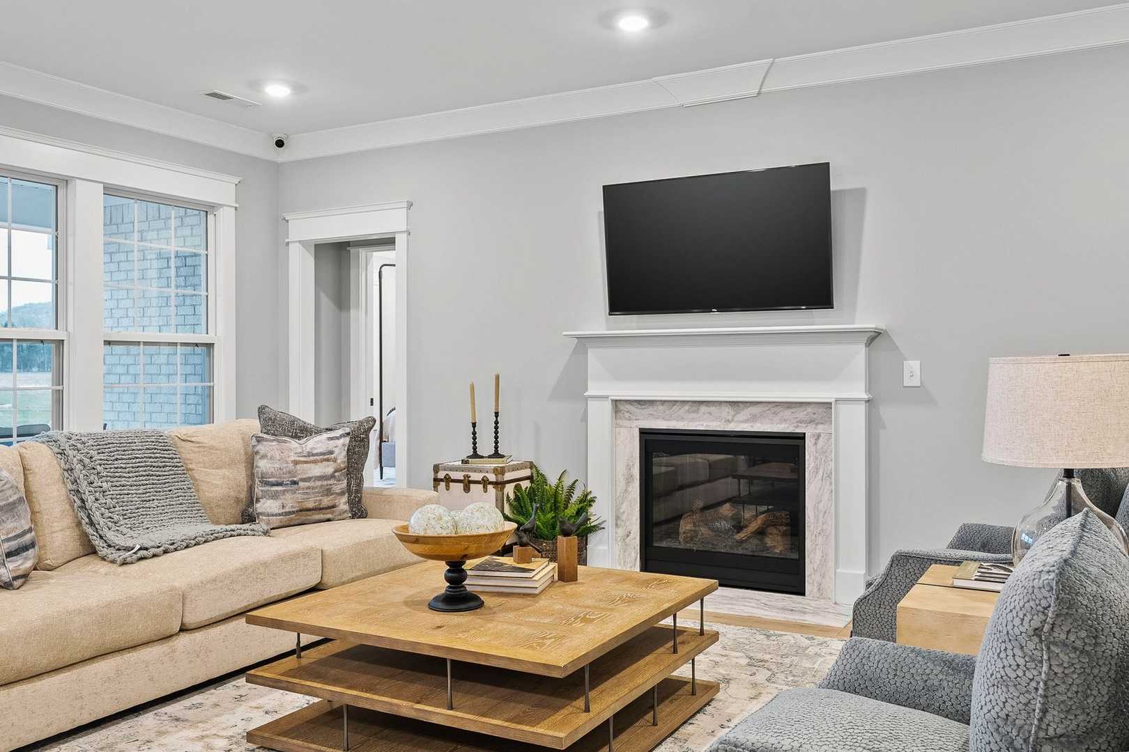 Cozy living room in The Oxford home with beige sofa, gas fireplace, mounted TV, wooden coffee table, and large windows