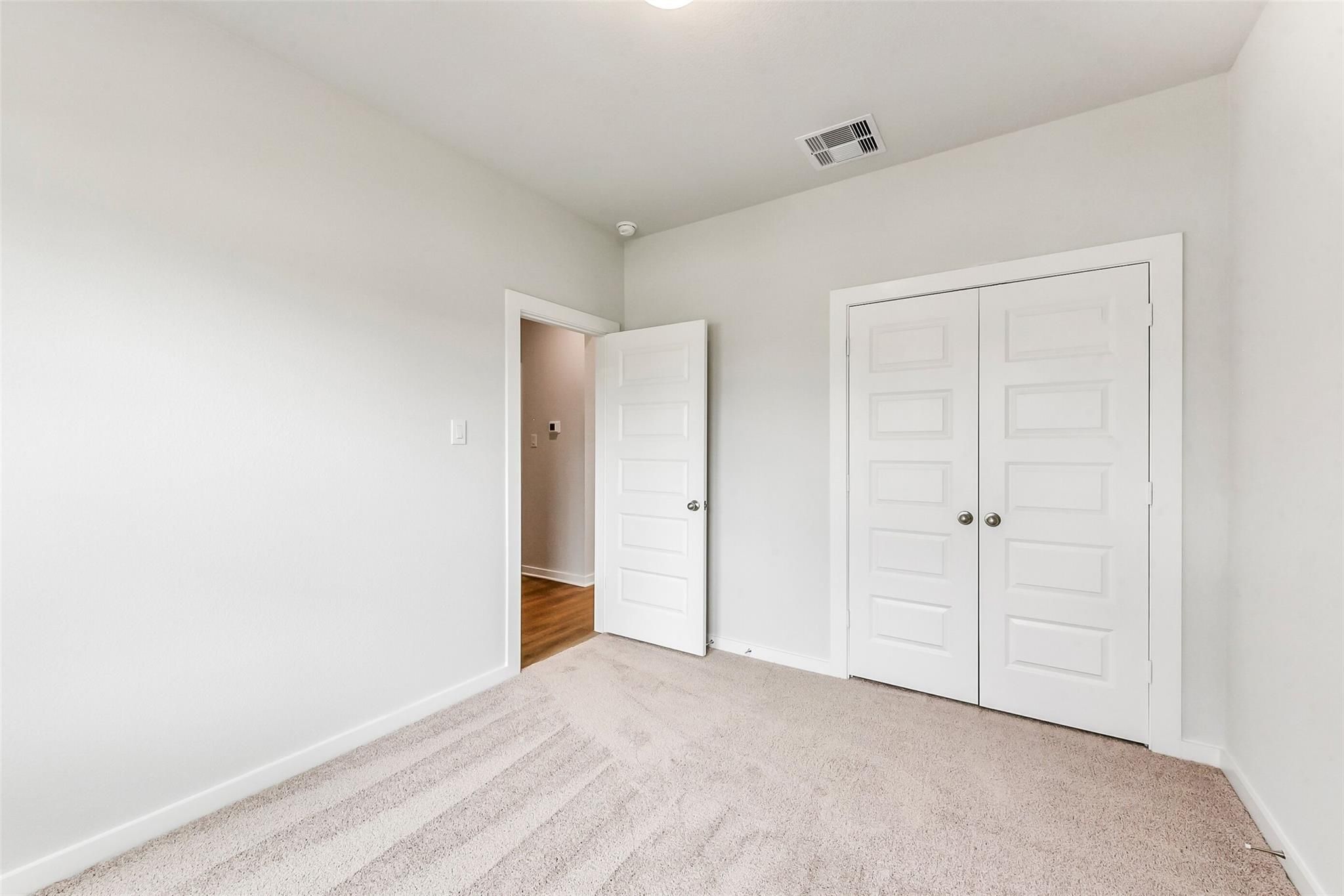 Bright bedroom with beige carpet, light gray walls, white double closet doors, and adjacent bath entry in Davidson Homes The Costa B, Dayton Texas