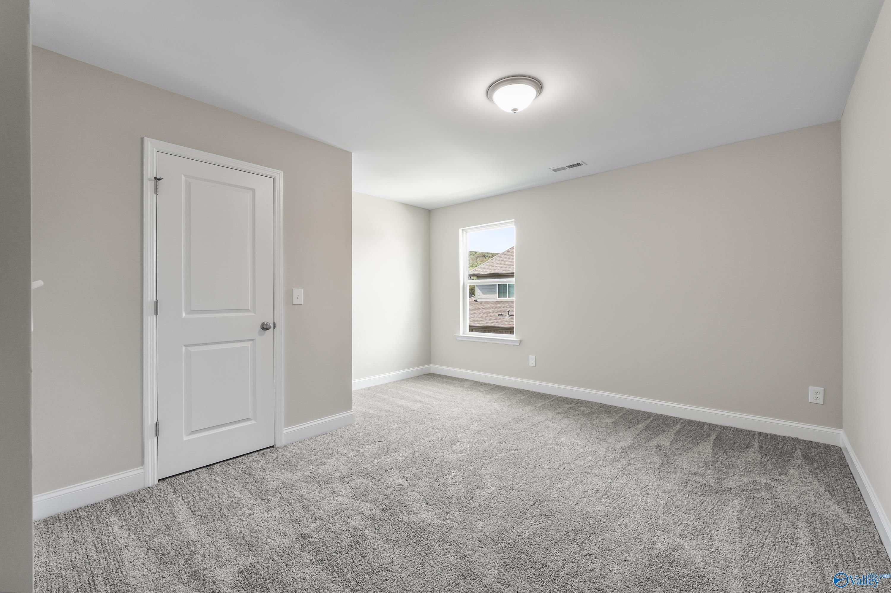 Empty secondary bedroom with neutral gray walls, white door, window view, and carpeted floor in Evermore Homes The Haven, Owens Cross Roads, Alabama