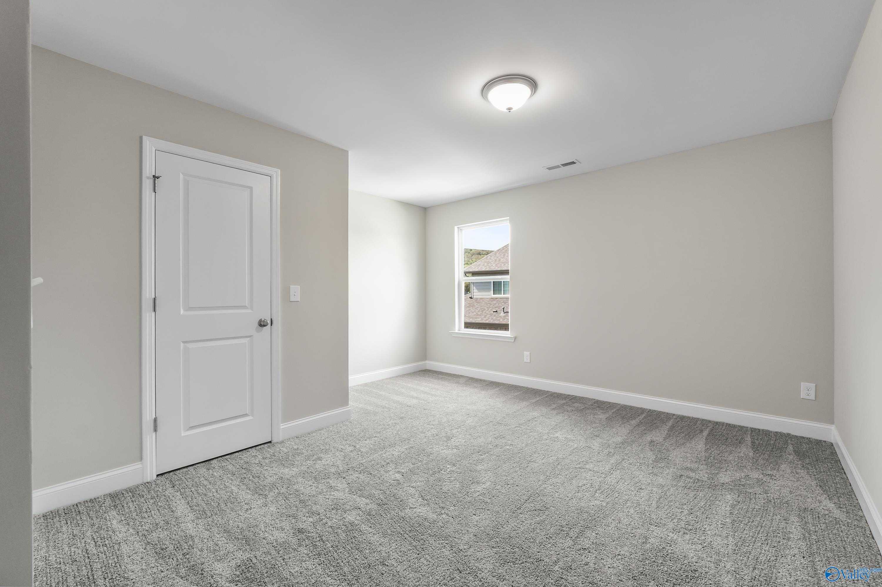 Empty secondary bedroom with neutral gray walls, white door, window view, and carpeted floor in Evermore Homes The Haven, Owens Cross Roads, Alabama