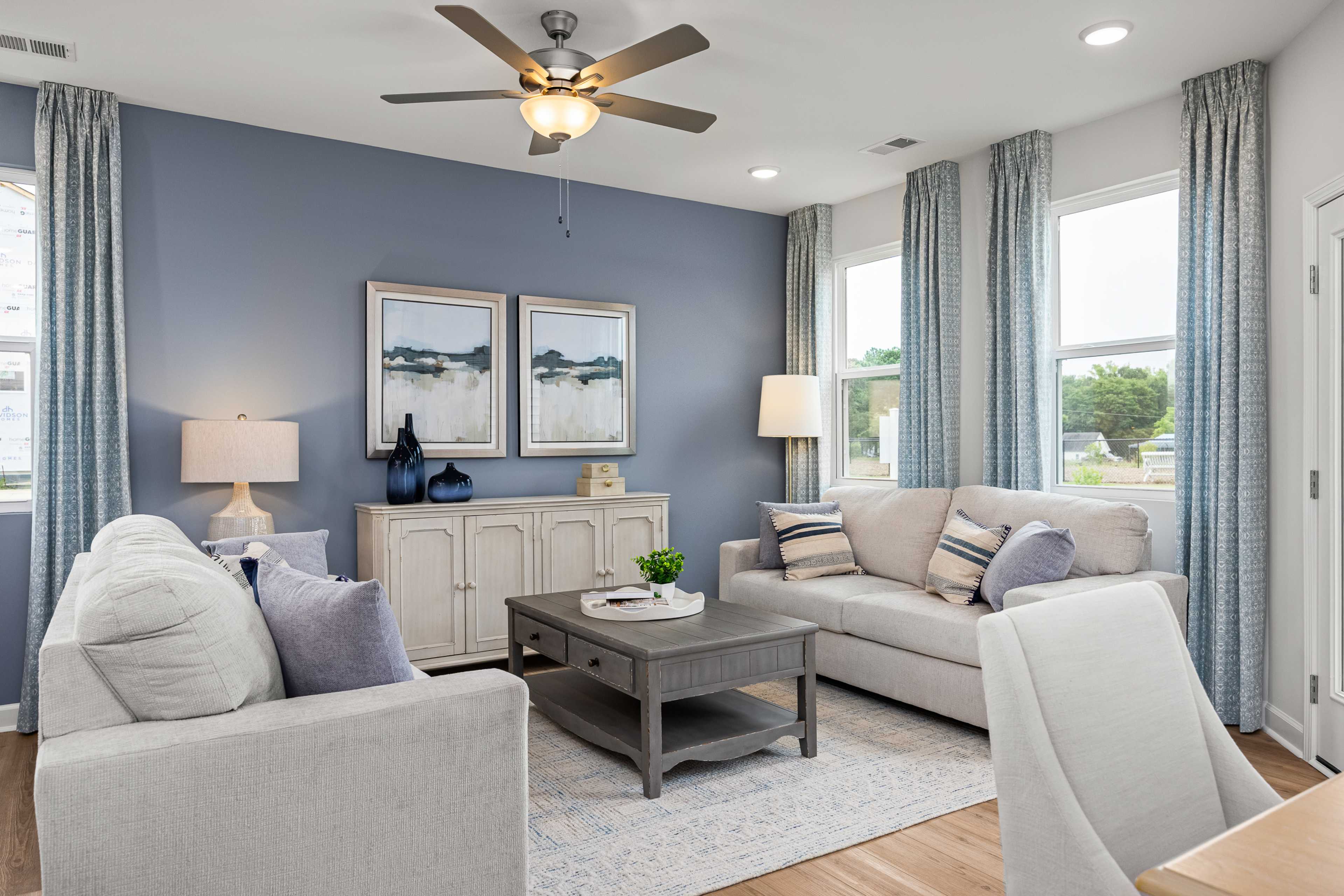 Cozy living room in Springvale Fuquay-Varina NC with blue walls white sofas hardwood floors and ceiling fan by Davidson Homes
