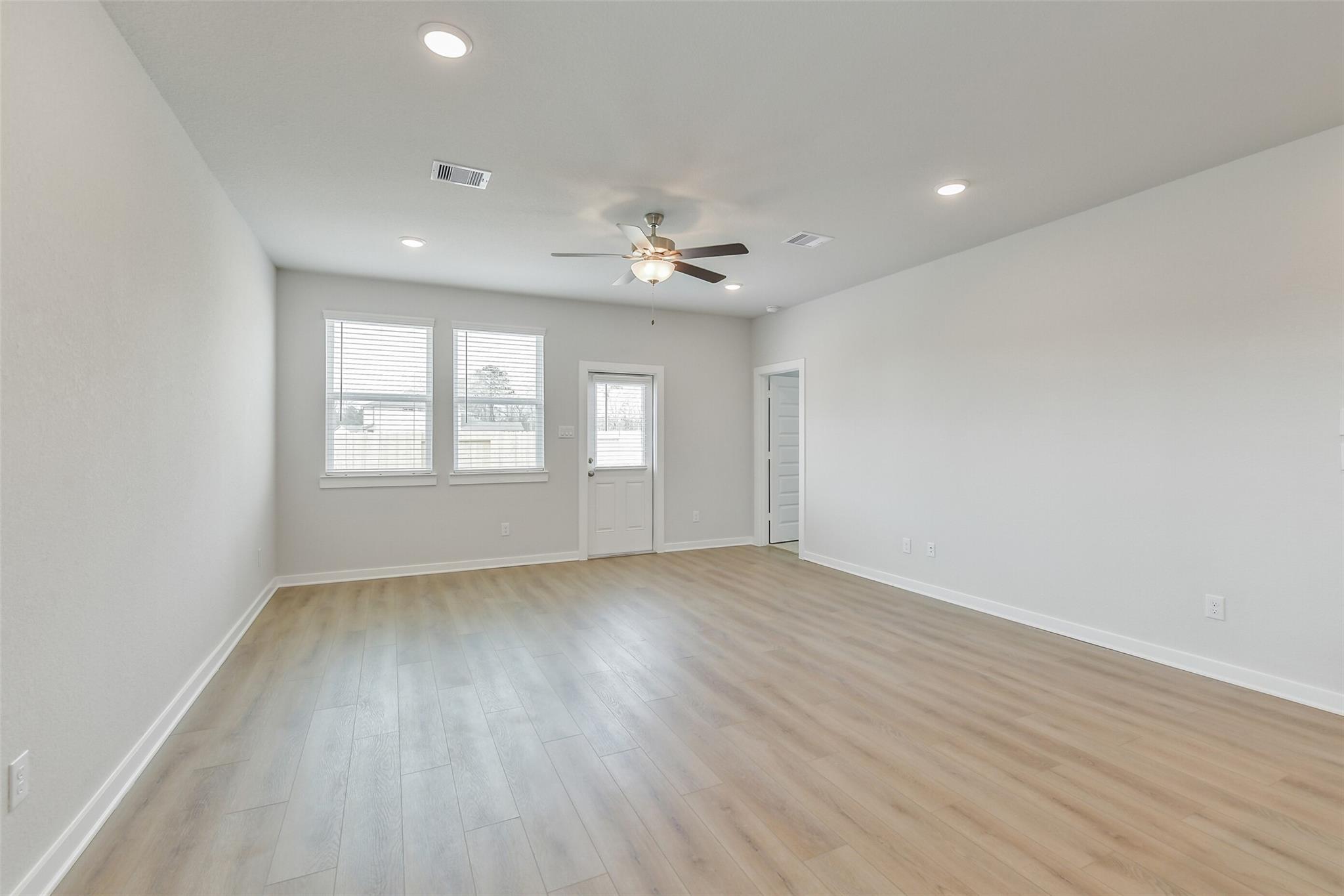 Bright living room with light gray walls, blonde wood floors, ceiling fan, recessed lights, and large windows in Davidson Homes The Brazos E, Cleveland, Texas