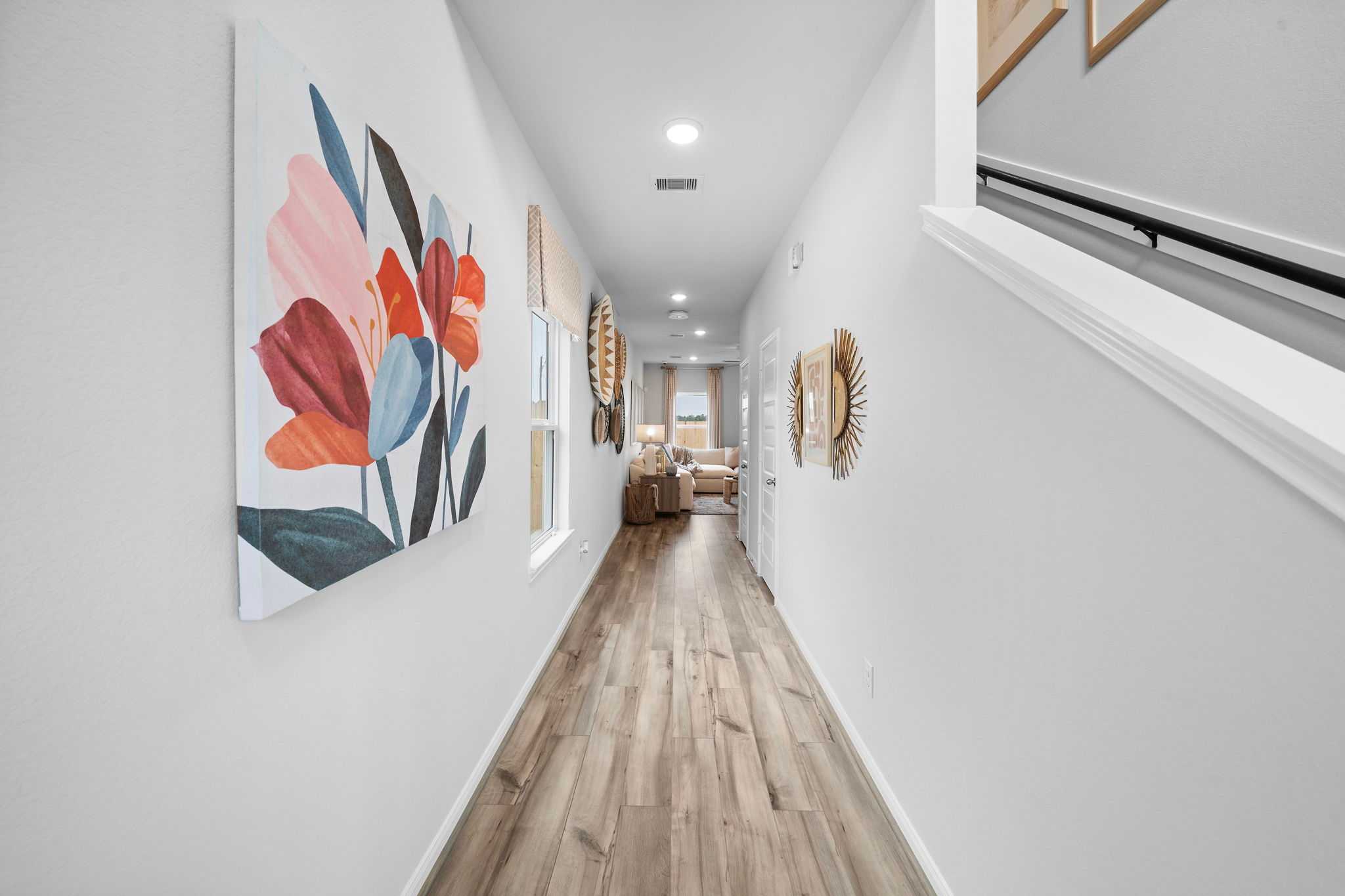 Spacious hallway with light hardwood floors, white walls, abstract floral wall art, and staircase at Hill & Dale Ranch in Splendora, Texas