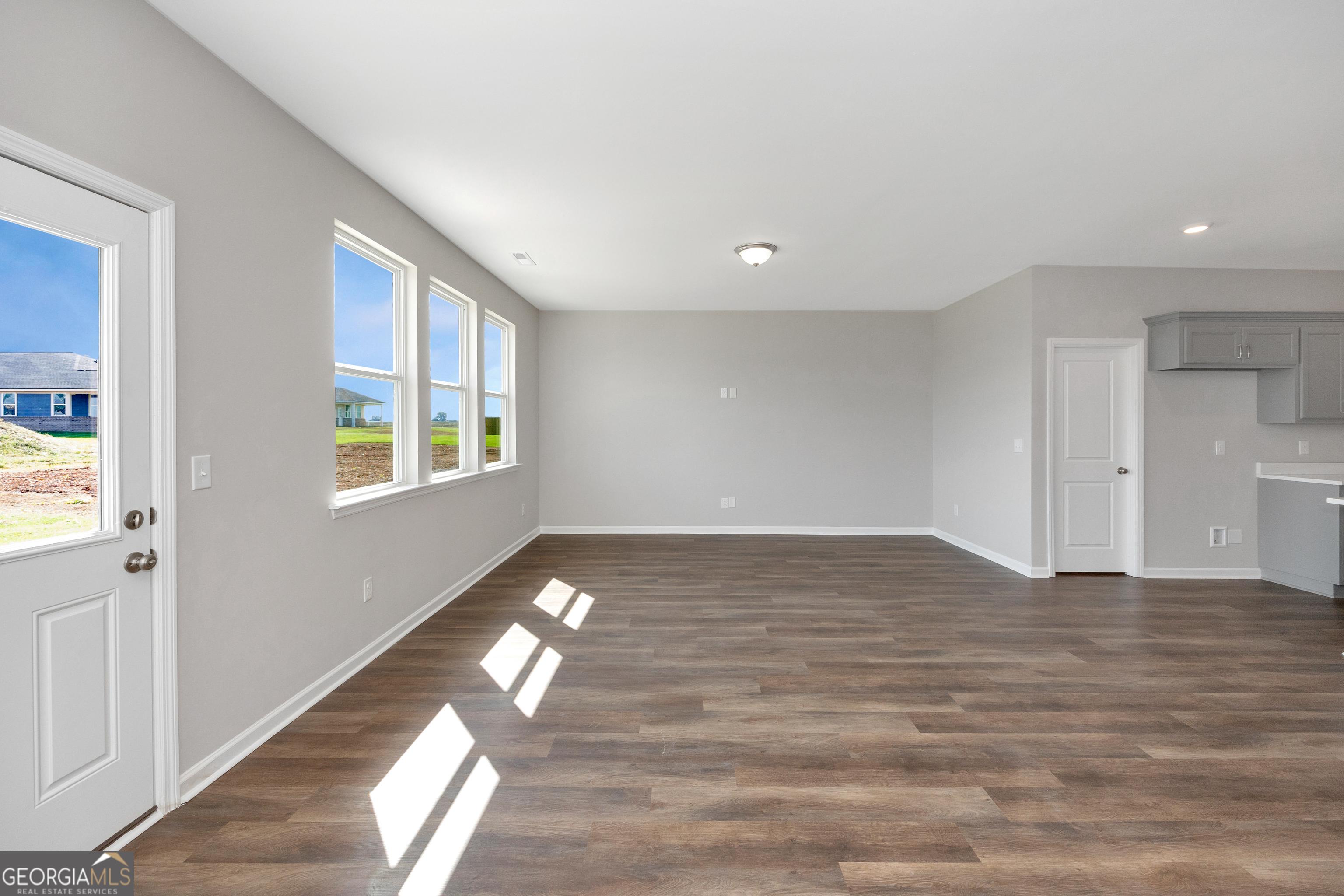 Sunlit empty living room with gray walls, large windows, hardwood floors, and open kitchen in The Luna floor plan, Ivy Glen, Perry, Georgia