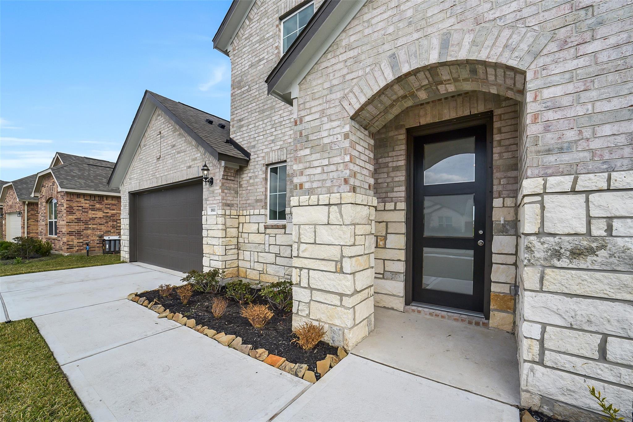 Modern two-story brick home front with arched entry, black glass door, stone pillars, 2-car garage, and landscaped walkway in Sierra Vista, Rosharon, Texas