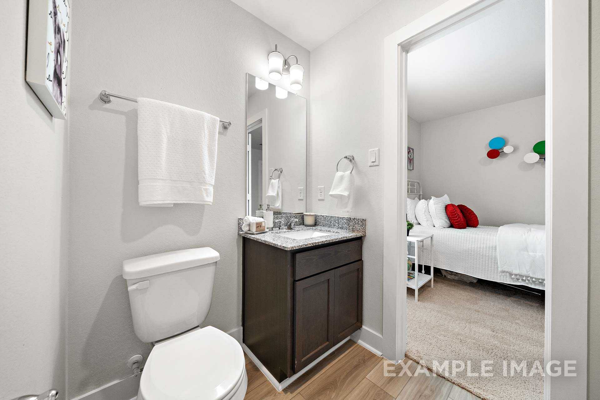 Master bathroom in The Lily A showcasing dark wood vanity, white fixtures, and adjacent bedroom with red pillows