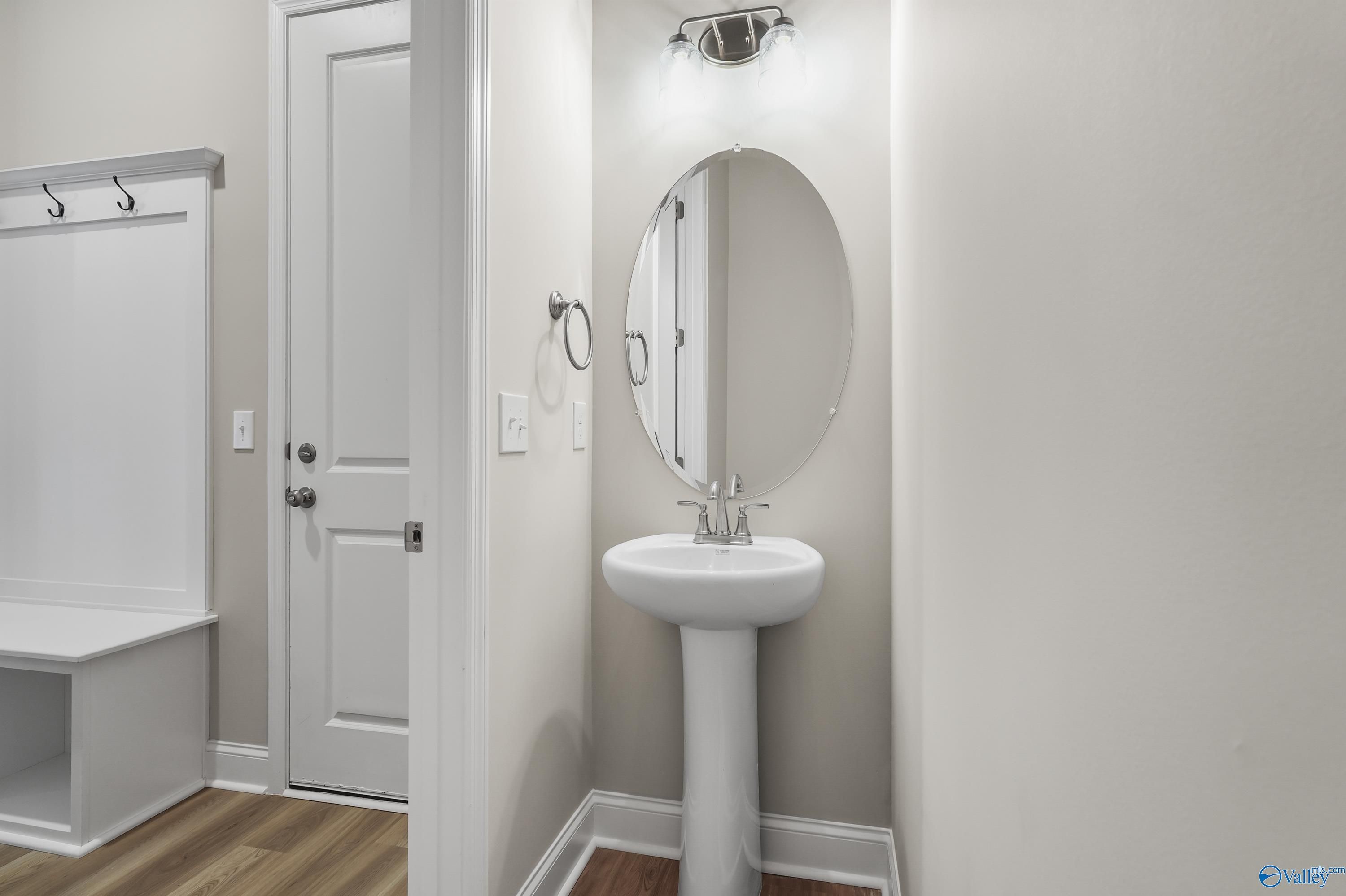 Elegant powder room with white pedestal sink, round mirror, coat hooks and bench in Evermore Homes The Haven, Owens Cross Roads, AL