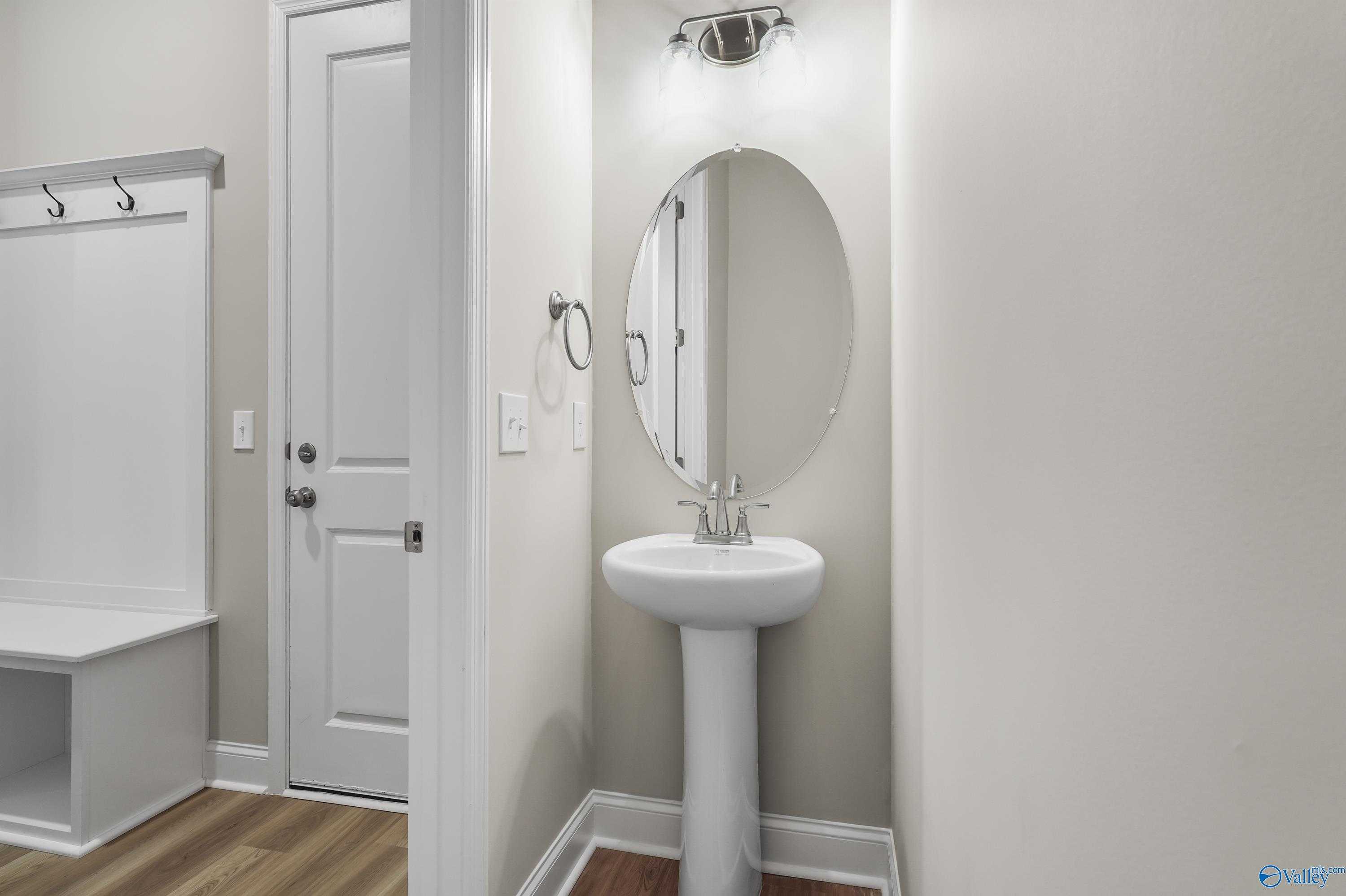 Elegant powder room with white pedestal sink, round mirror, coat hooks and bench in Evermore Homes The Haven, Owens Cross Roads, AL
