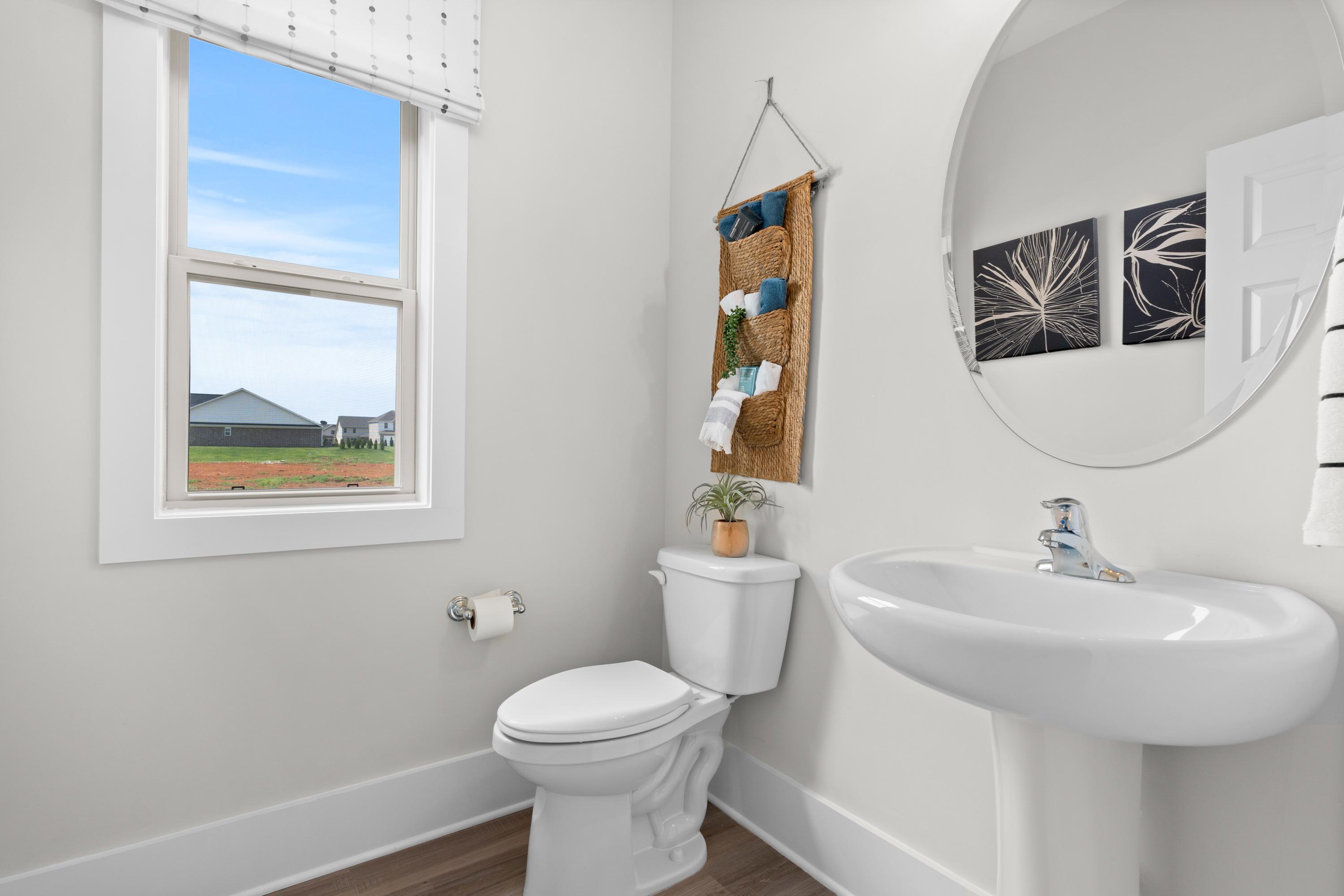 Bright powder room in The Chelsea A home featuring white pedestal sink, round mirror, toilet, macrame plant hanger, and window with scenic view