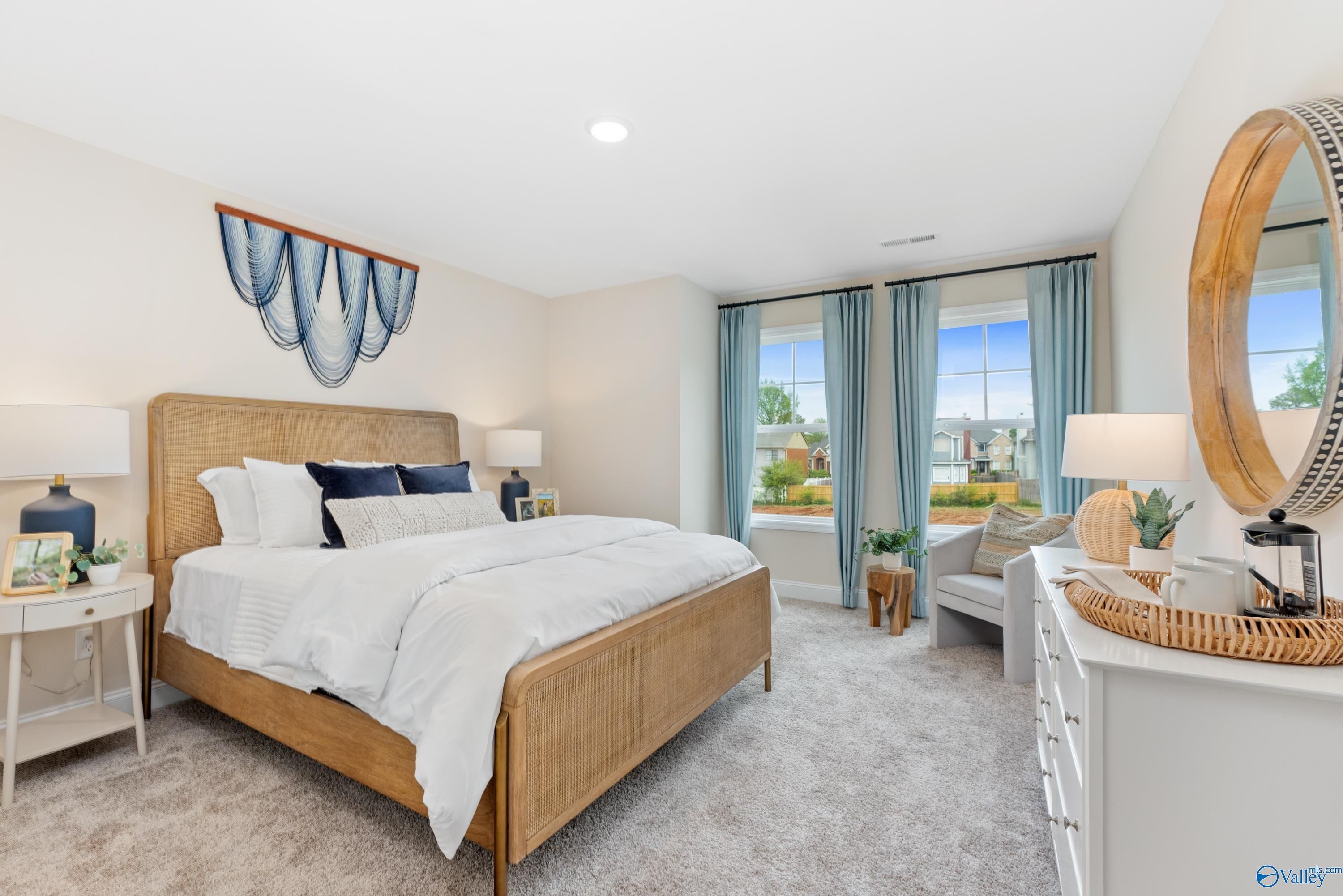 Serene bedroom with light wood bed, white bedding, blue pillows, large windows, round mirror in Davidson Homes The Camden, Huntsville