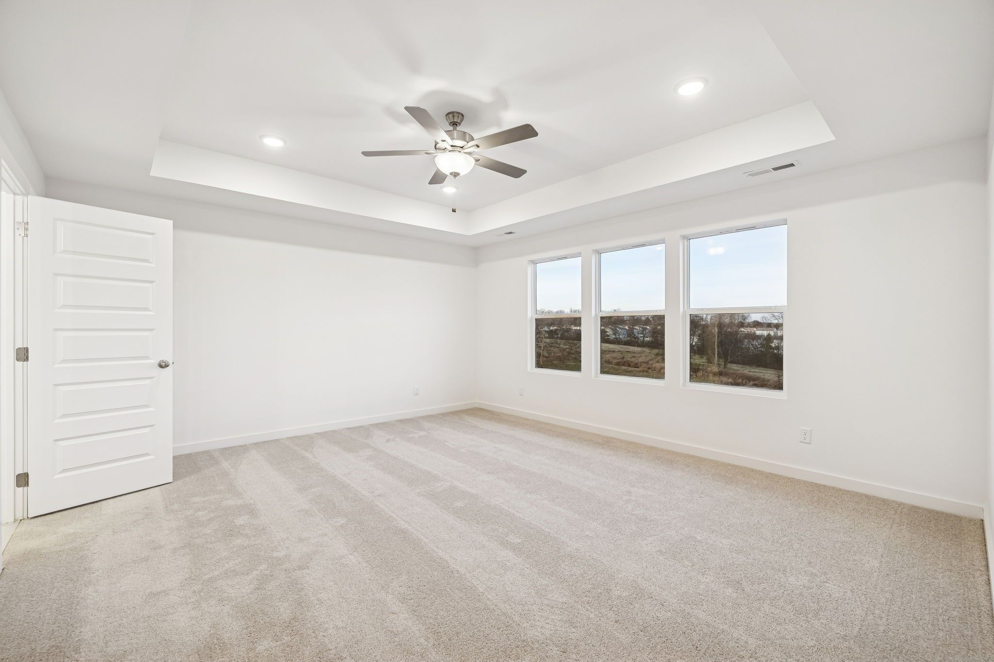Bright empty bedroom with tray ceiling, ceiling fan, large triple windows, and beige carpet in Davidson Homes The Willow, White House, TN