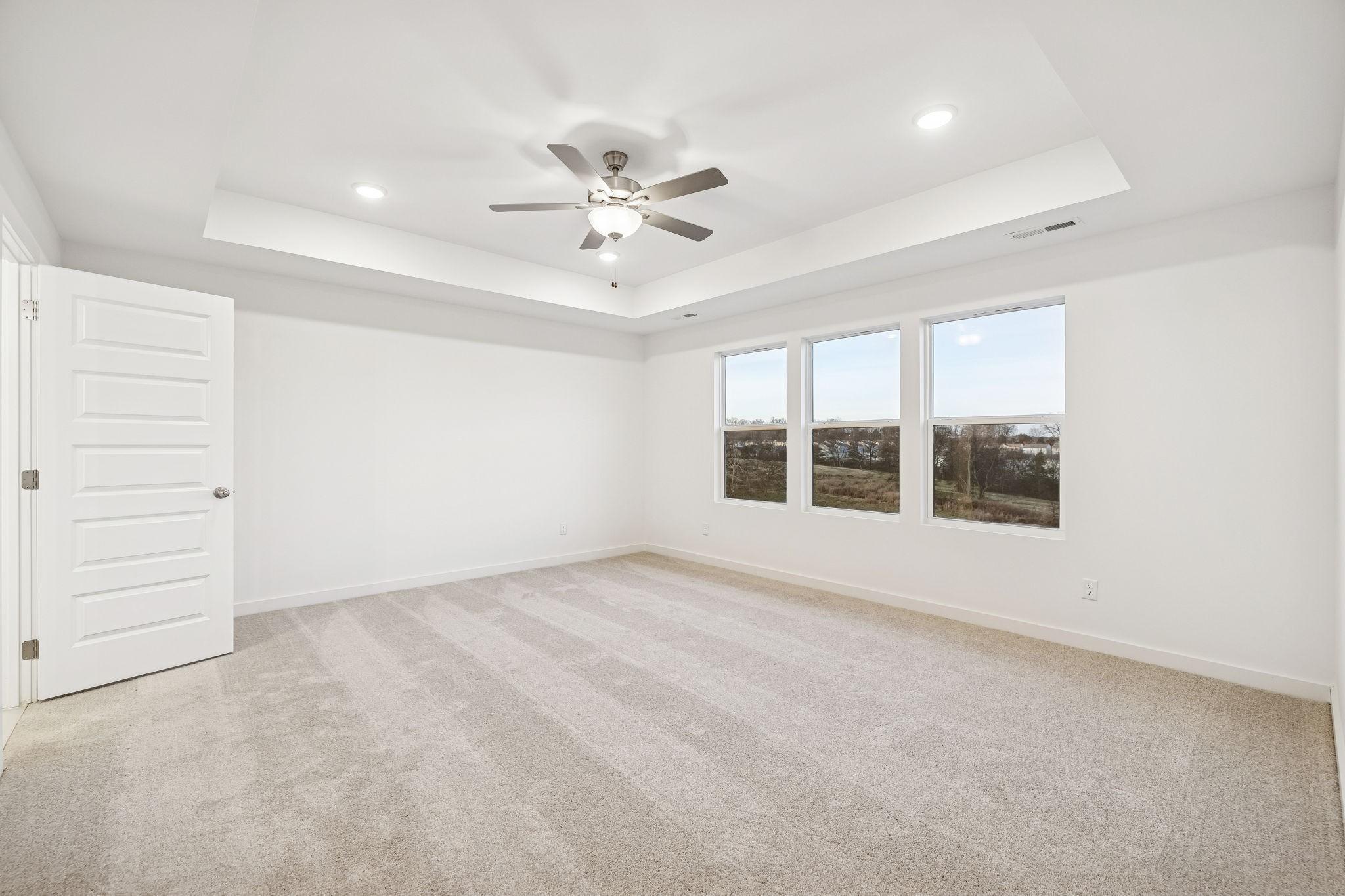Bright empty bedroom with tray ceiling, ceiling fan, large triple windows, and beige carpet in Davidson Homes The Willow, White House, TN