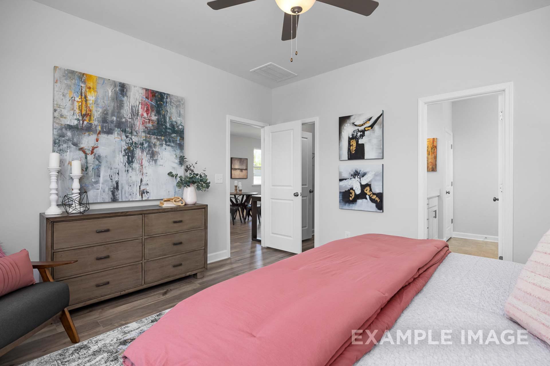 Spacious master bedroom in The Carter B featuring pink king bed, abstract wall art, wooden dresser, ceiling fan, and bathroom doorway