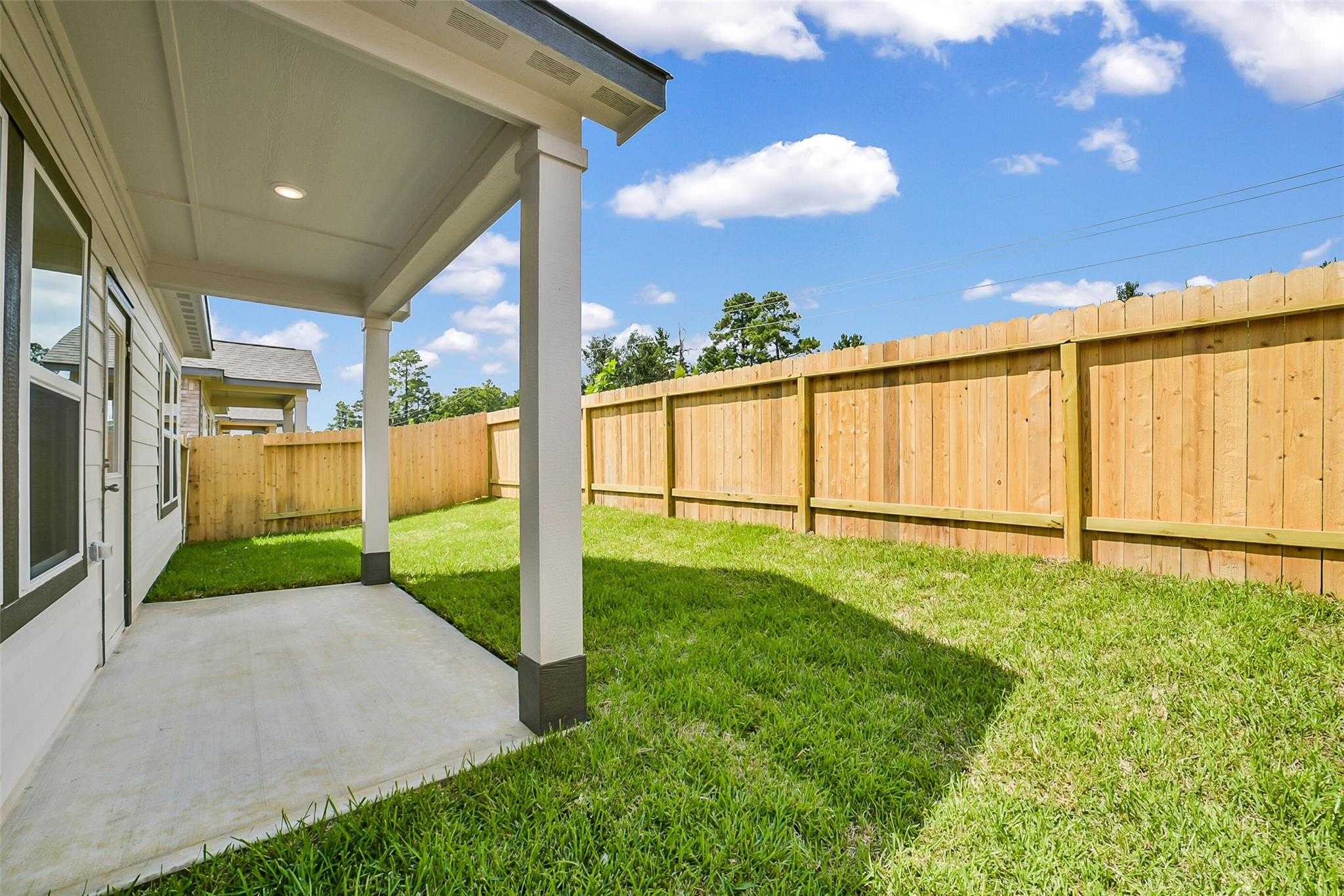 Covered patio with white columns, wooden privacy fence, and lush green lawn beside Davidson Homes The Blanco E in Magnolia, Texas