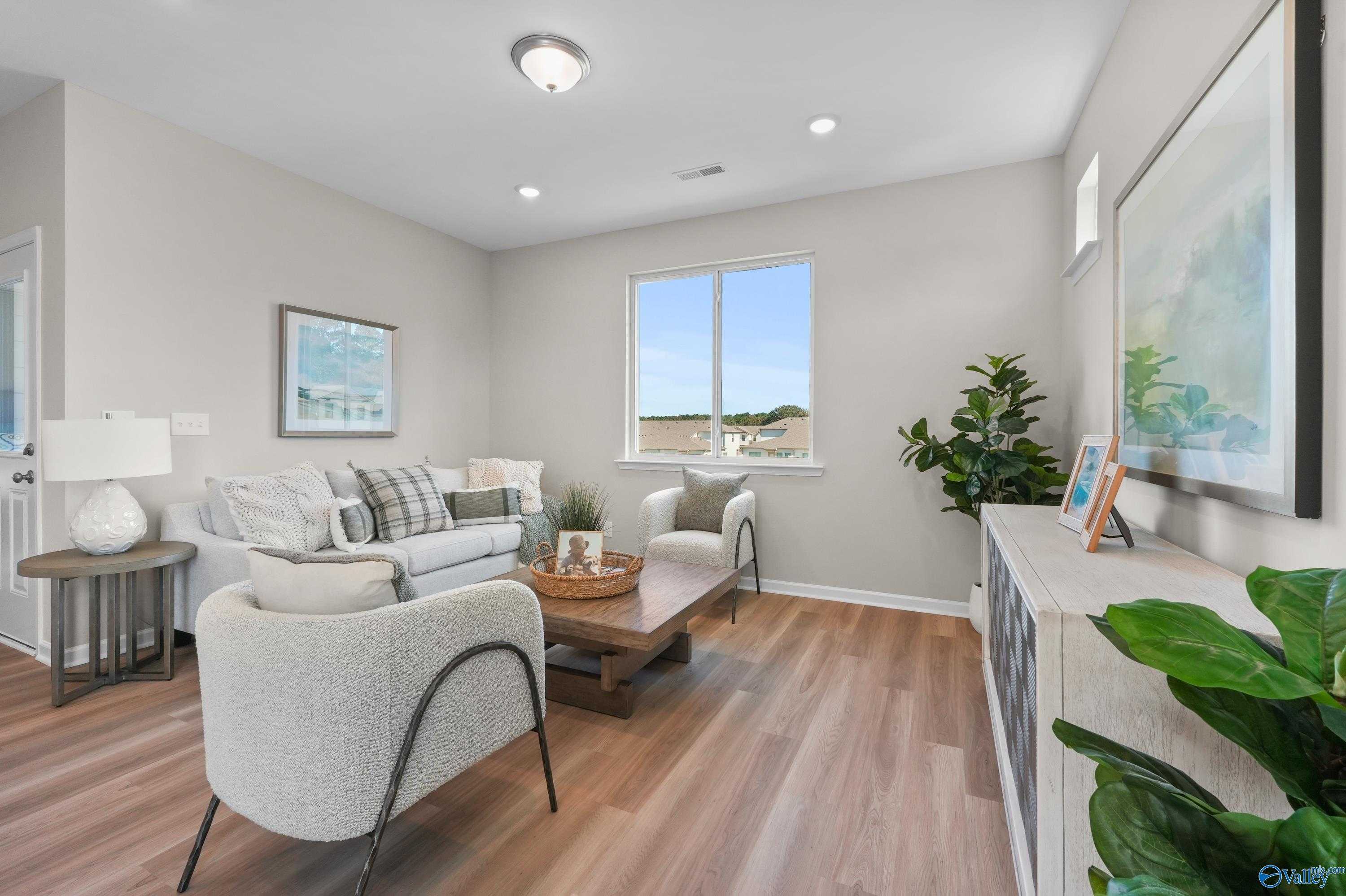 Bright living room with neutral walls, plush beige sofa, armchairs, hardwood floors, and plants in Evermore Homes The Malibu, Madison, Alabama