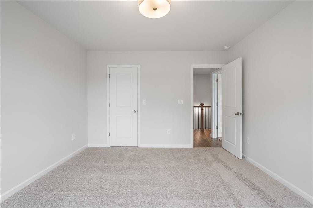 Empty secondary bedroom featuring neutral beige walls, plush carpet floor, closet door, and open hallway to staircase in Davidson Homes The Cary A, Kennesaw, GA
