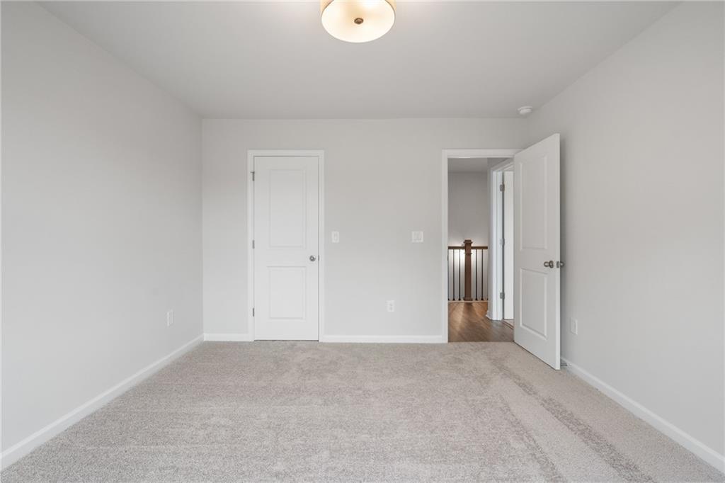 Spacious empty bedroom with neutral gray walls, beige carpet, white doors, and hallway stairs in Davidson Homes The Cary A, Kennesaw, GA