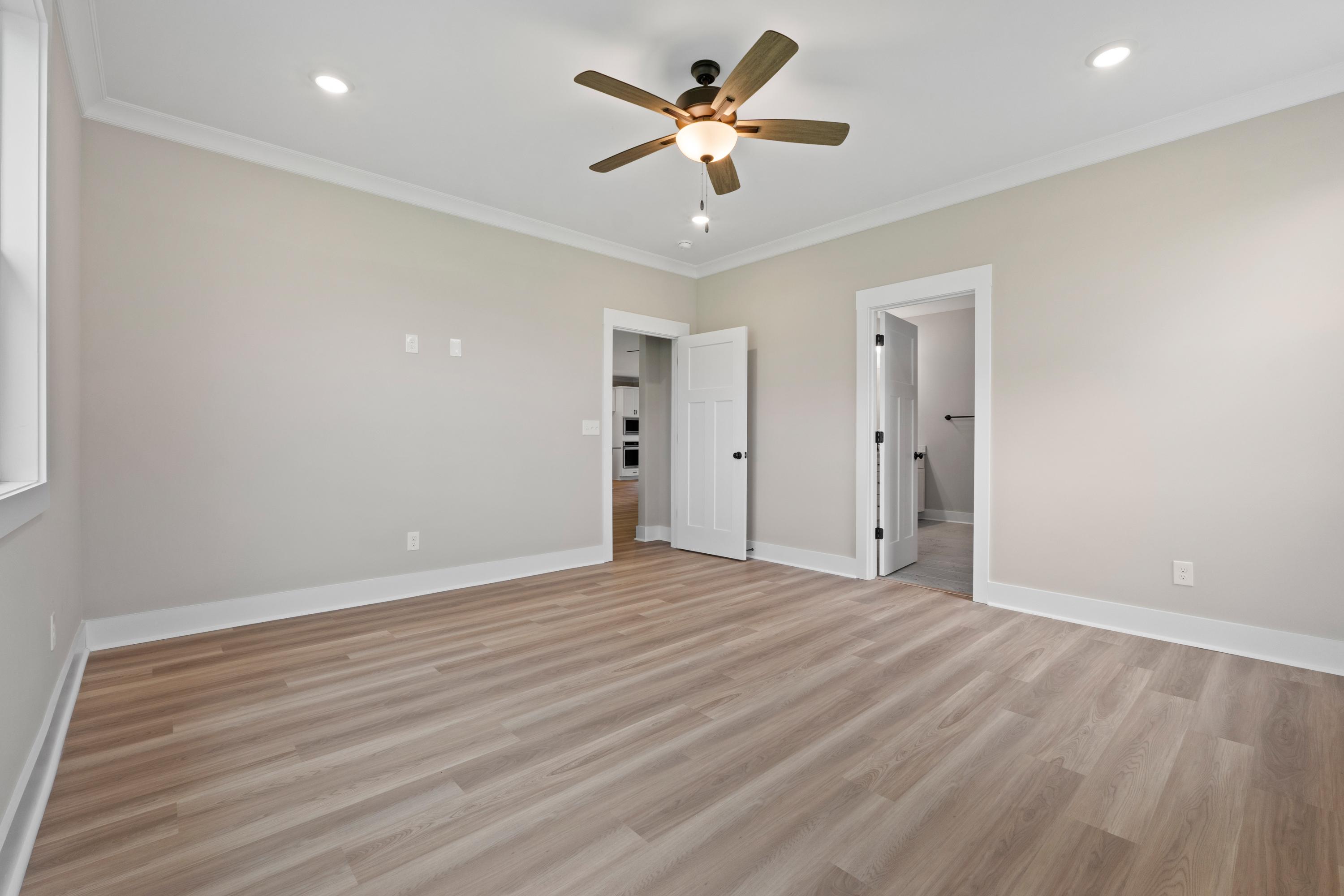 Spacious master bedroom in The Oxford B with light hardwood floors, beige walls, ceiling fan, and en-suite bath access