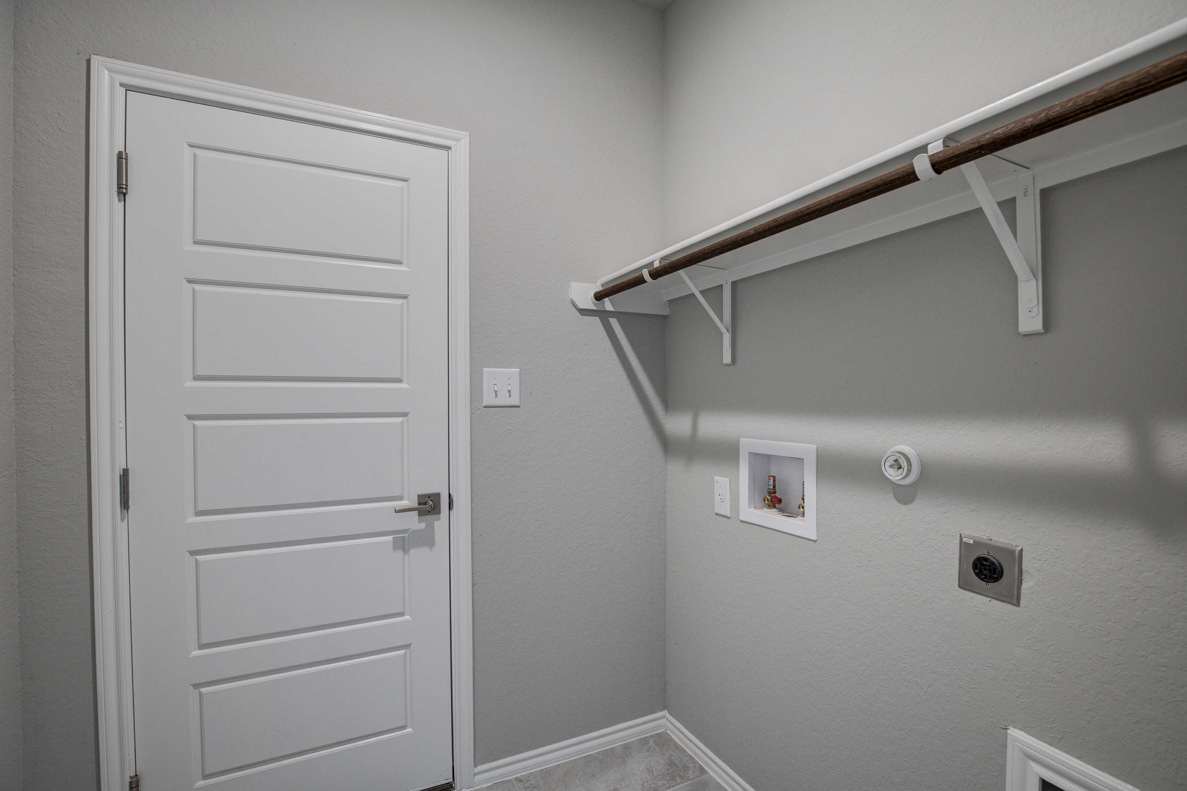 Spacious laundry room in The Frio B home with white paneled door, wooden open shelves, utility sink, and gray walls