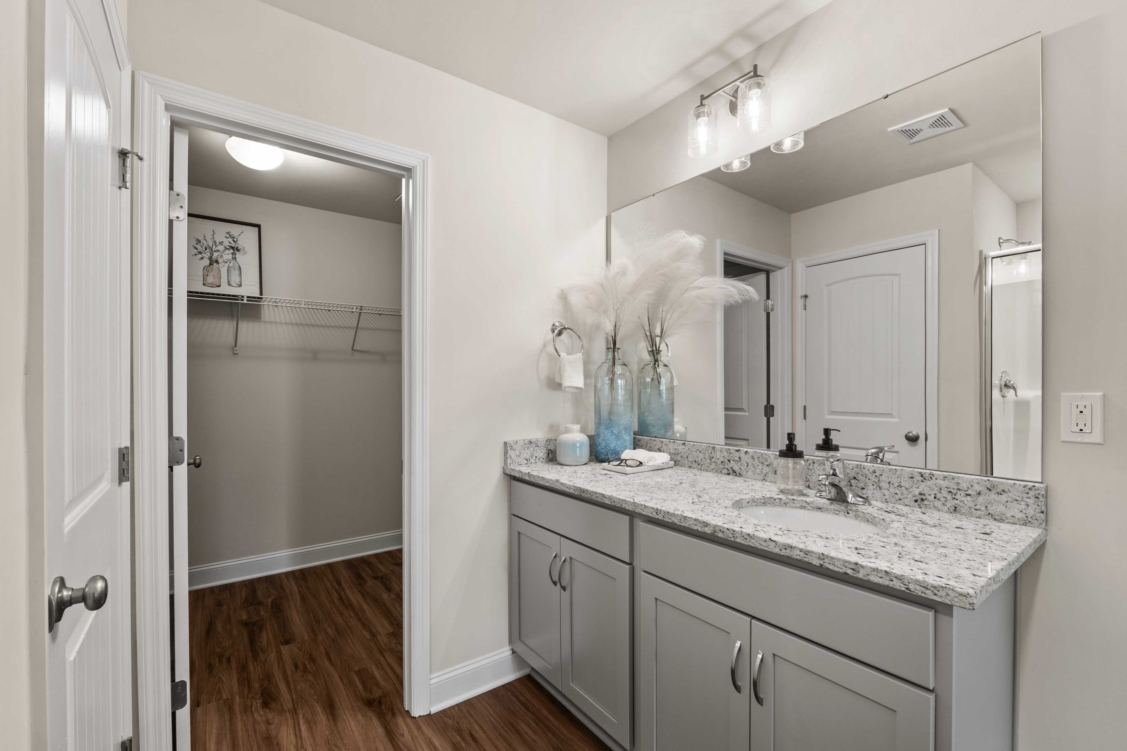 Elegant bathroom vanity at Ramsay Cove in Owens Cross Roads Alabama with gray cabinets, quartz countertop, blue vases, and open walk-in closet