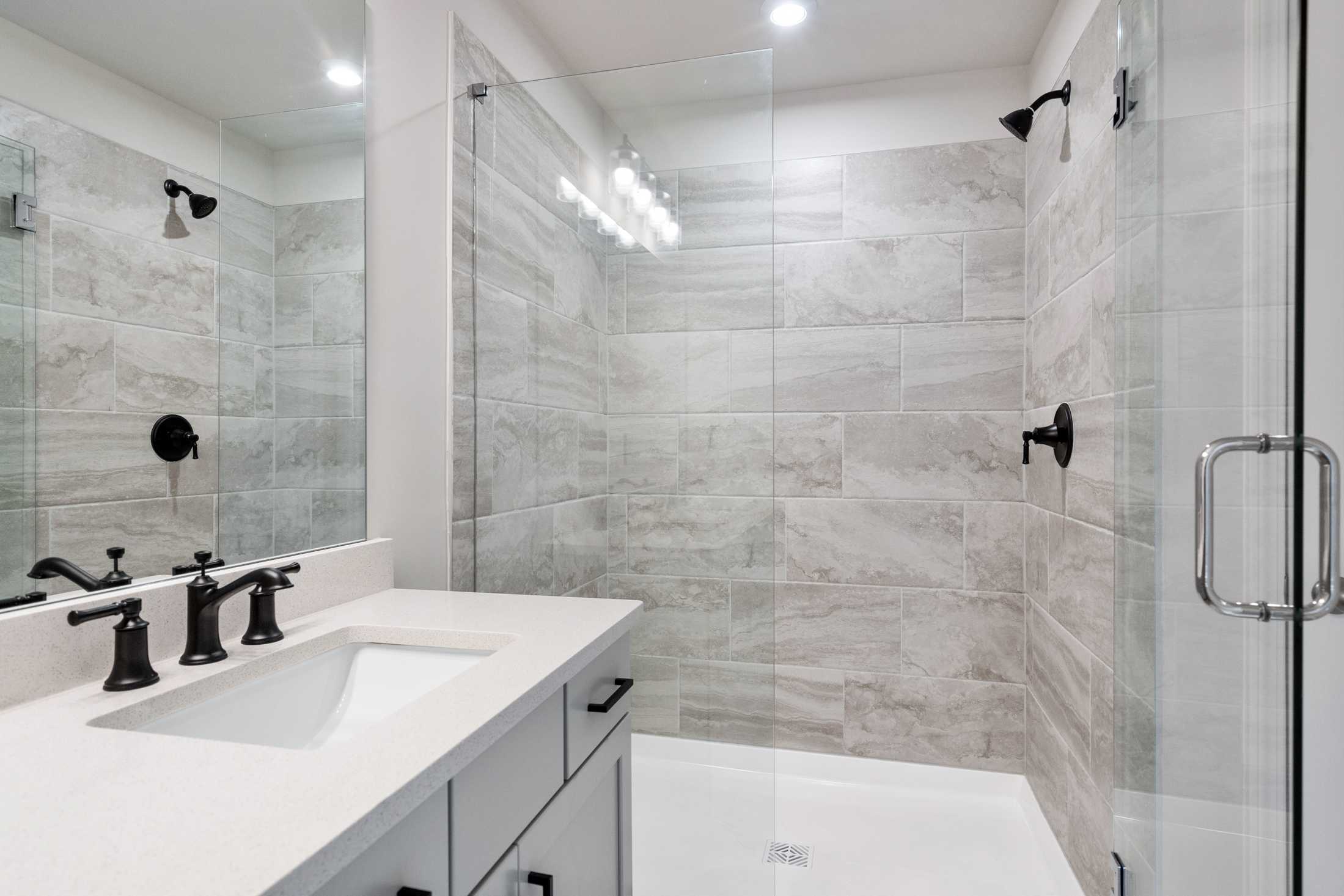 Modern master bathroom in The Cary B featuring gray tiled shower, frameless glass enclosure, and double vanity with black fixtures