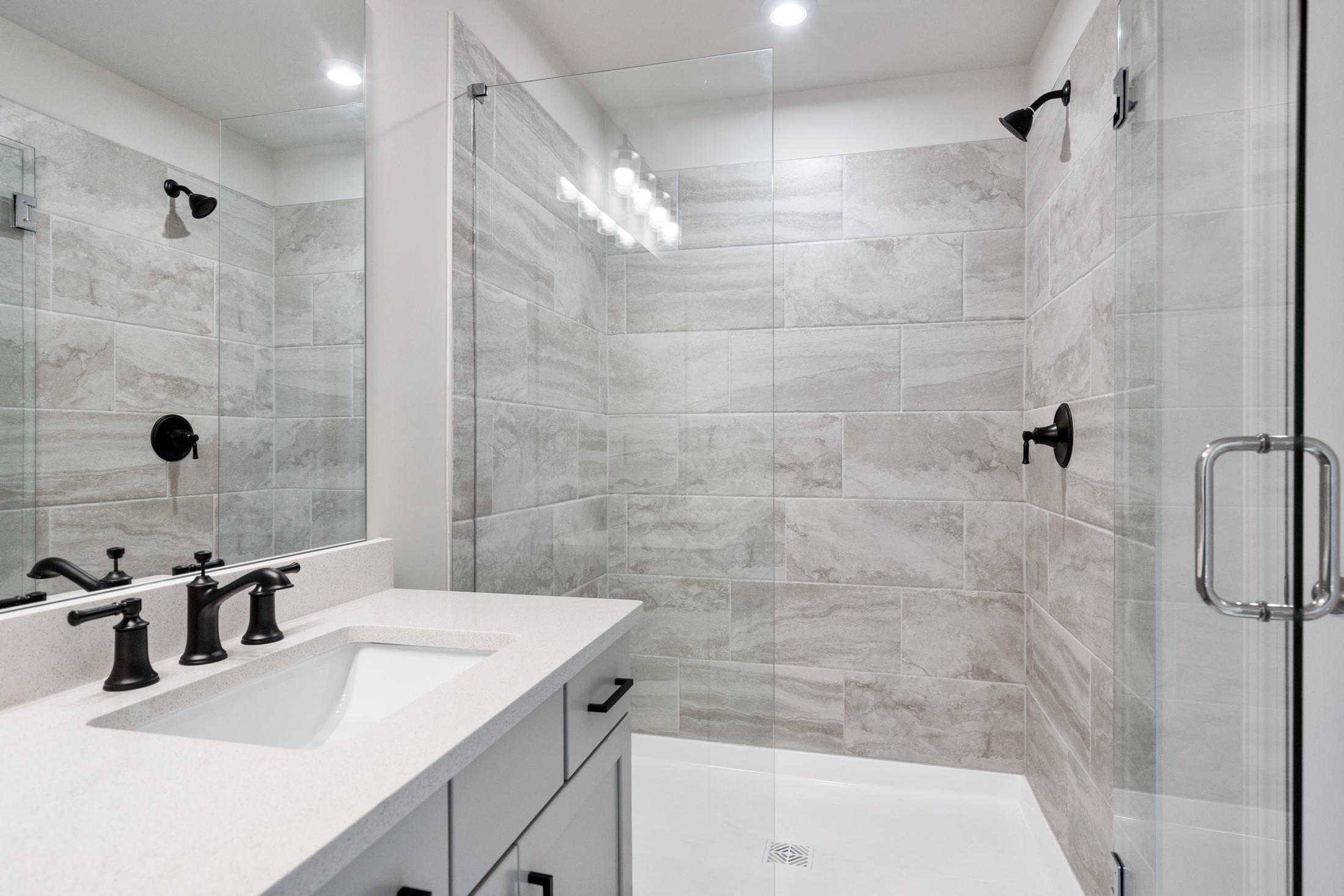 Modern master bathroom in The Cary B featuring gray tiled shower, frameless glass enclosure, and double vanity with black fixtures