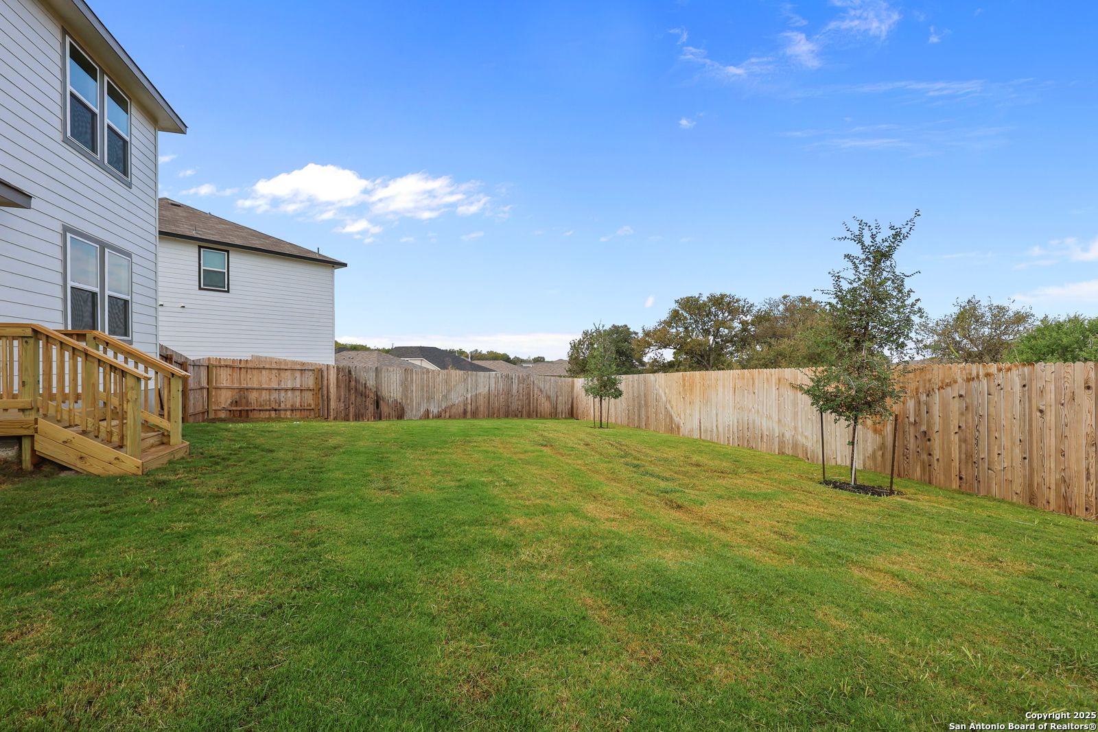 Spacious fenced backyard with lush green lawn, wooden deck stairs, and young trees in Davidson Homes The Murray J, Royal Crest, San Antonio, Texas