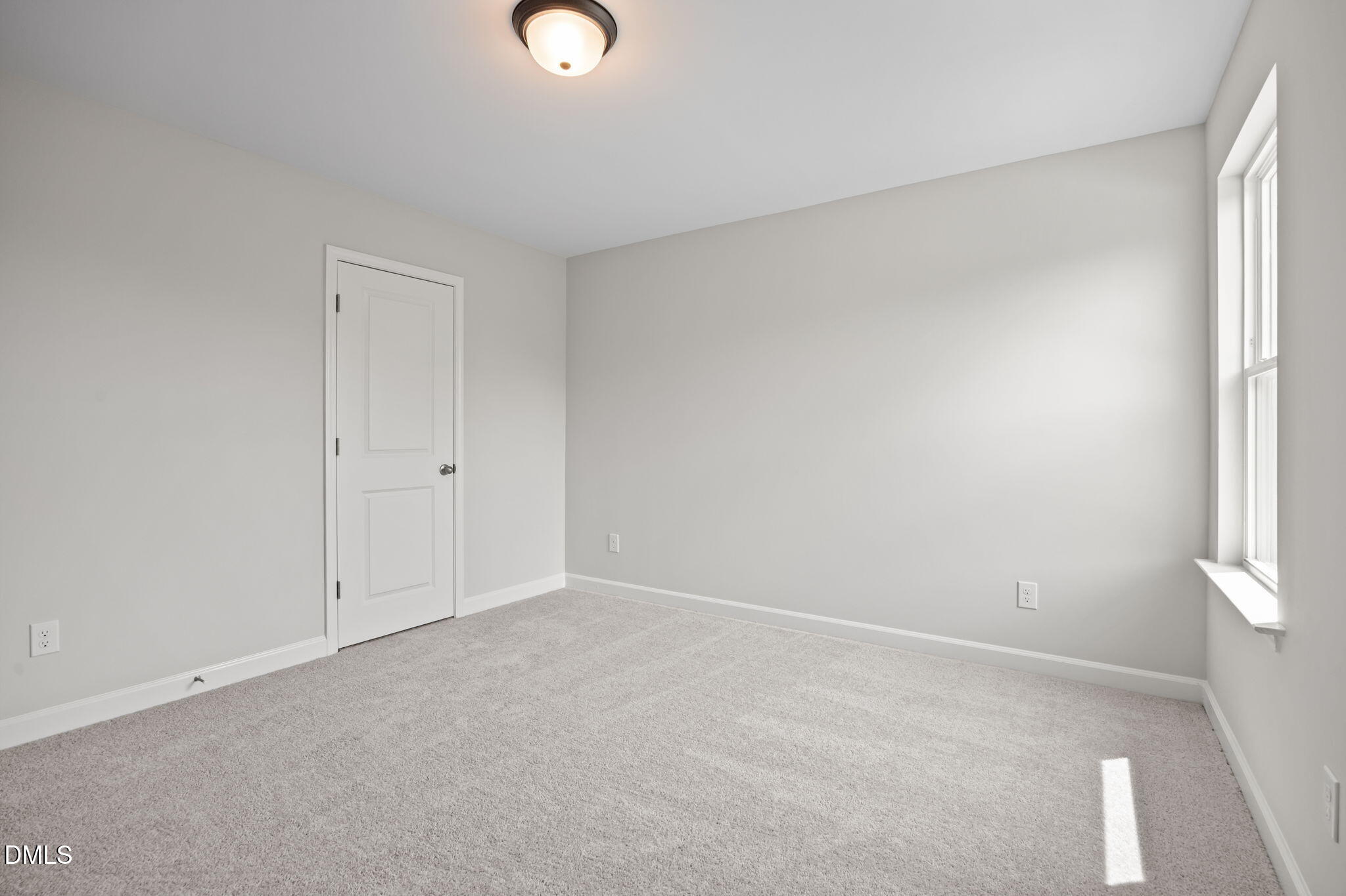 Bright empty secondary bedroom with light gray walls, white door, window sill, ceiling light, and plush carpet in The Willow D, Wendell, NC