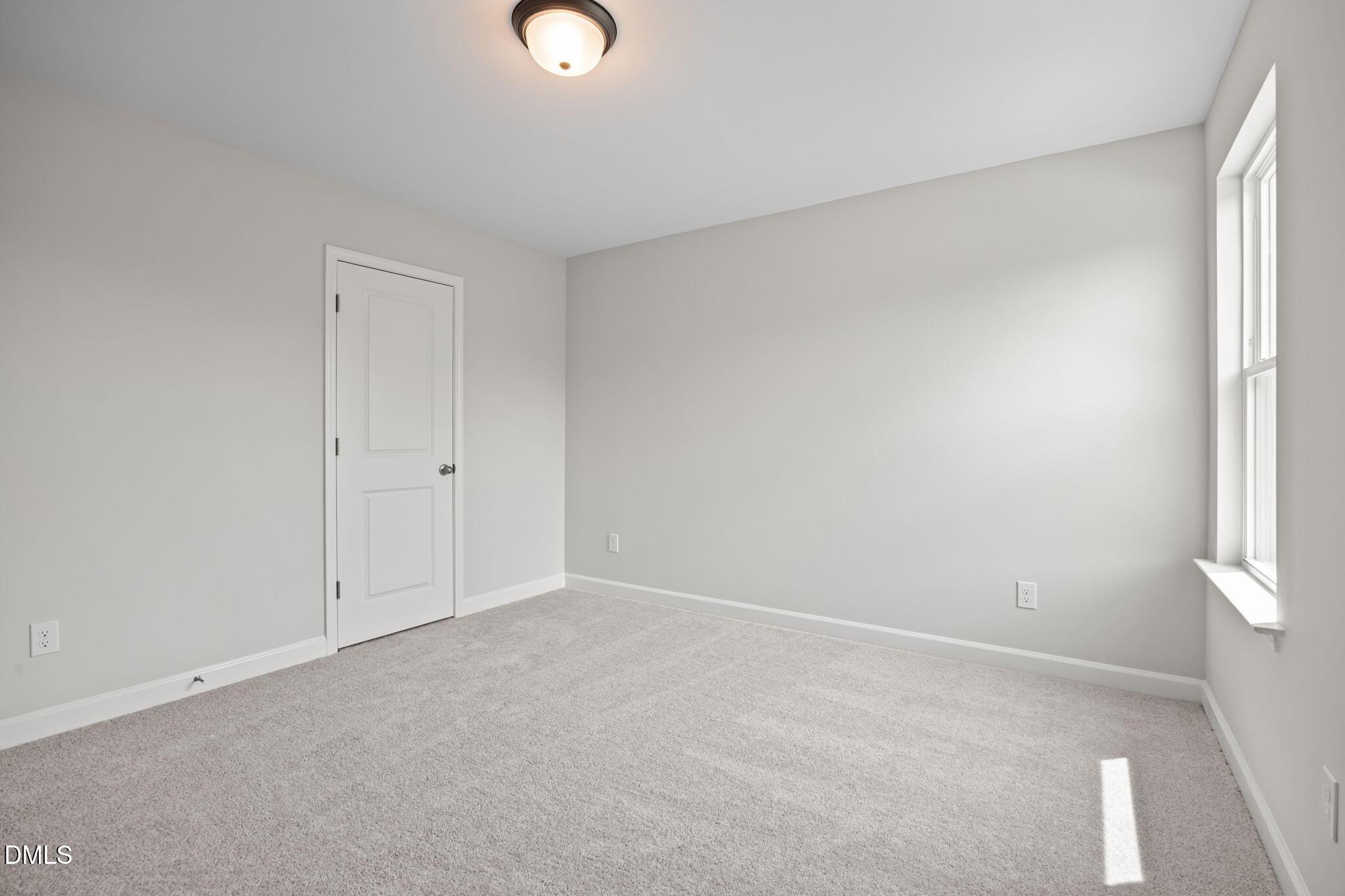Bright empty secondary bedroom with light gray walls, white door, window sill, ceiling light, and plush carpet in The Willow D, Wendell, NC