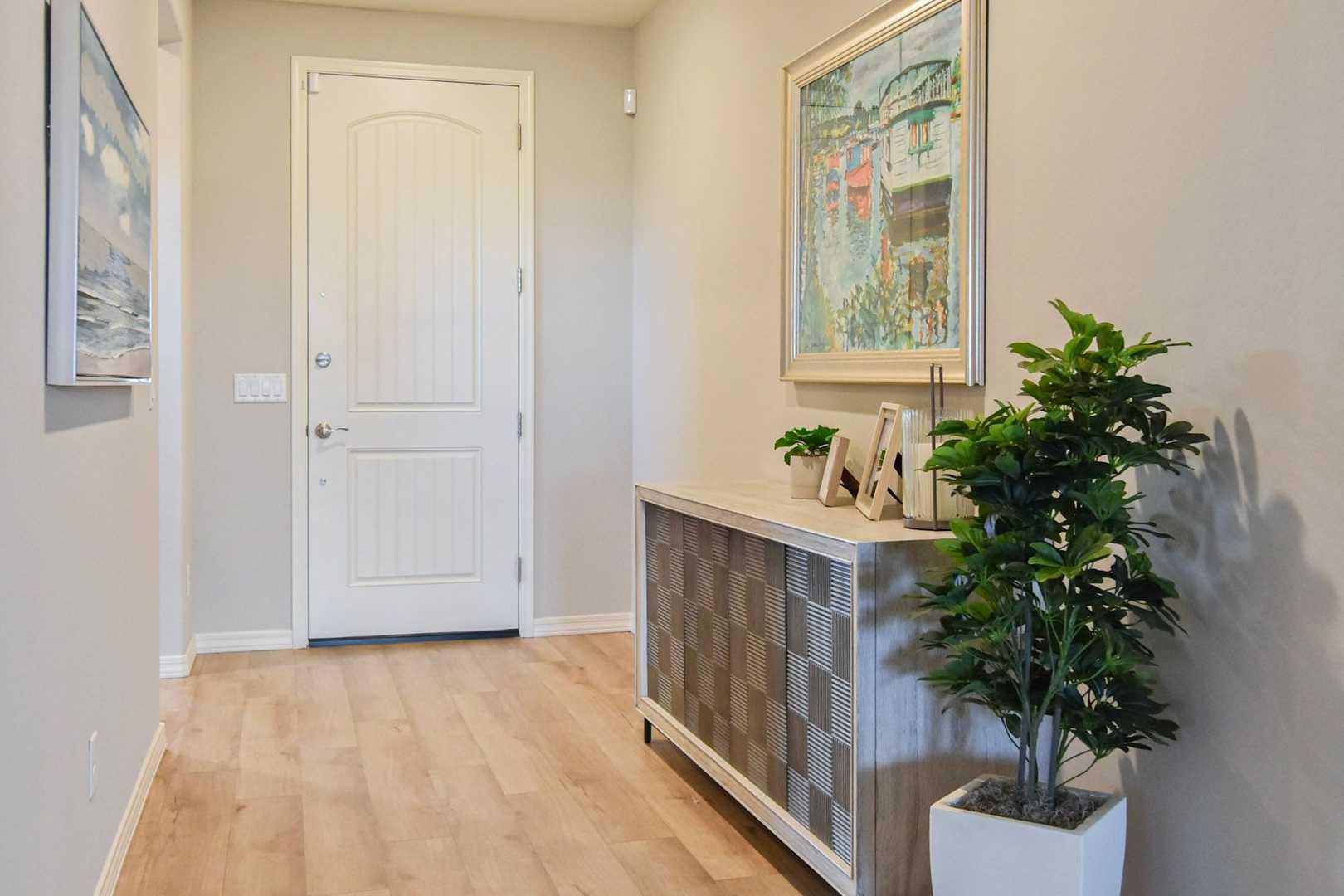 Spacious entryway in The Harmony home design featuring white paneled door, gray walls, hardwood floors, console table with potted plant and seascape artwork