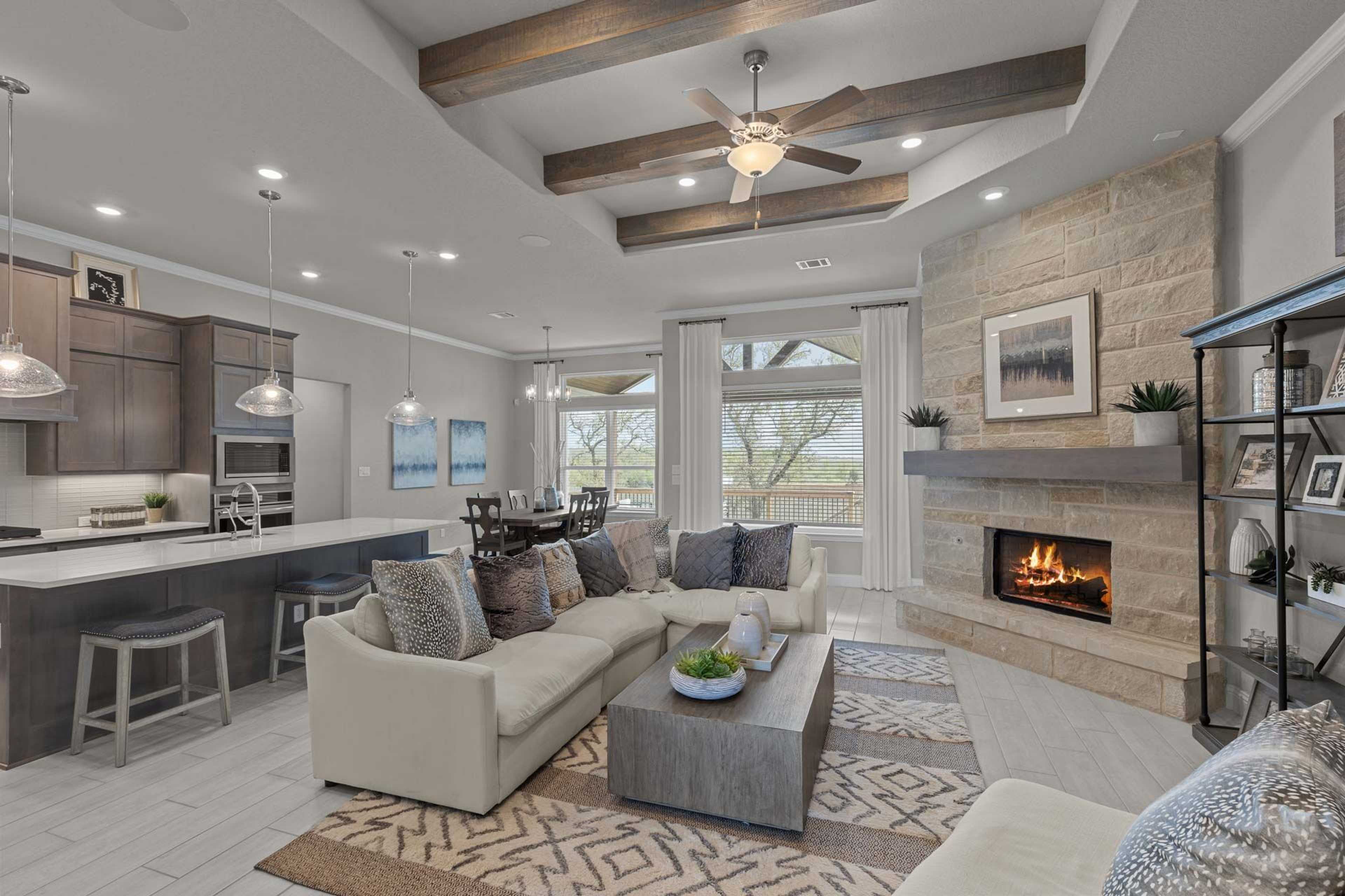 Open-concept living kitchen in Ladera San Antonio TX home by Davidson Homes featuring exposed beams, stone fireplace, beige sofa, and bar island