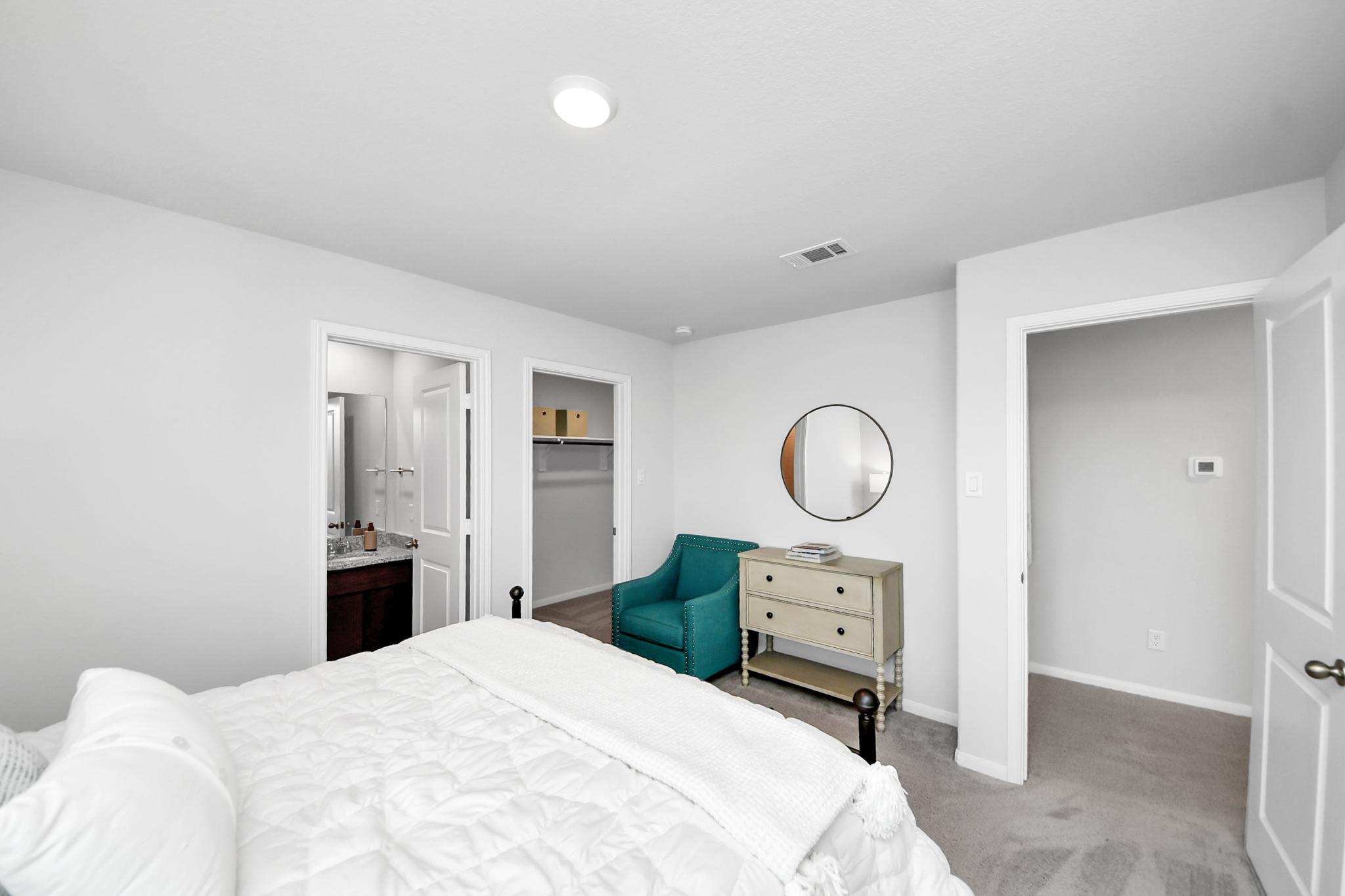 Spacious bedroom at Sunterra in Katy, TX by Davidson Homes featuring a white bed, teal accent chair, and en-suite bathroom.