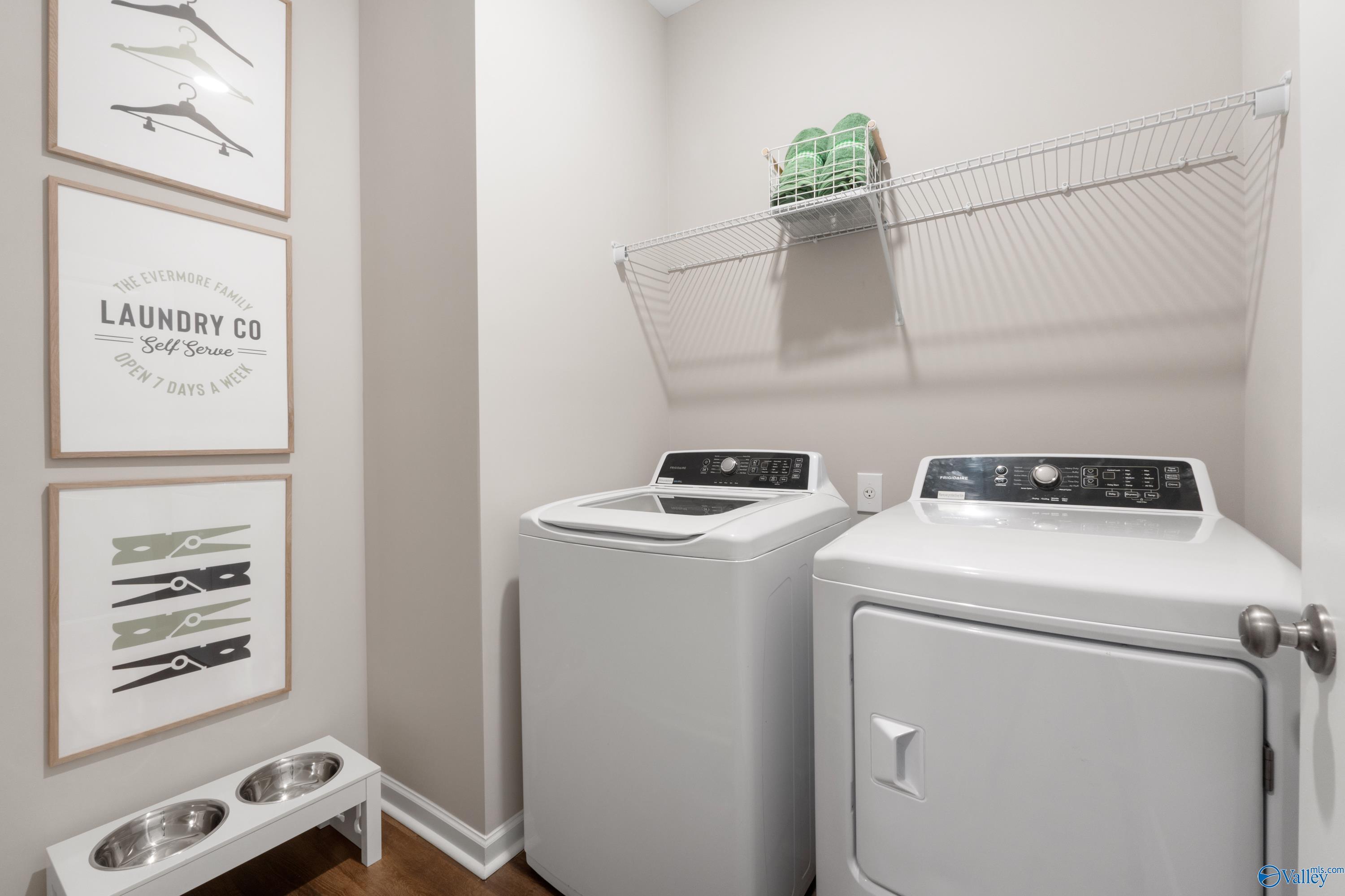 Functional laundry room with white washer-dryer set, wire shelving, green towels, laundry art, pet bowls in Davidson Homes The Stella, Madison AL