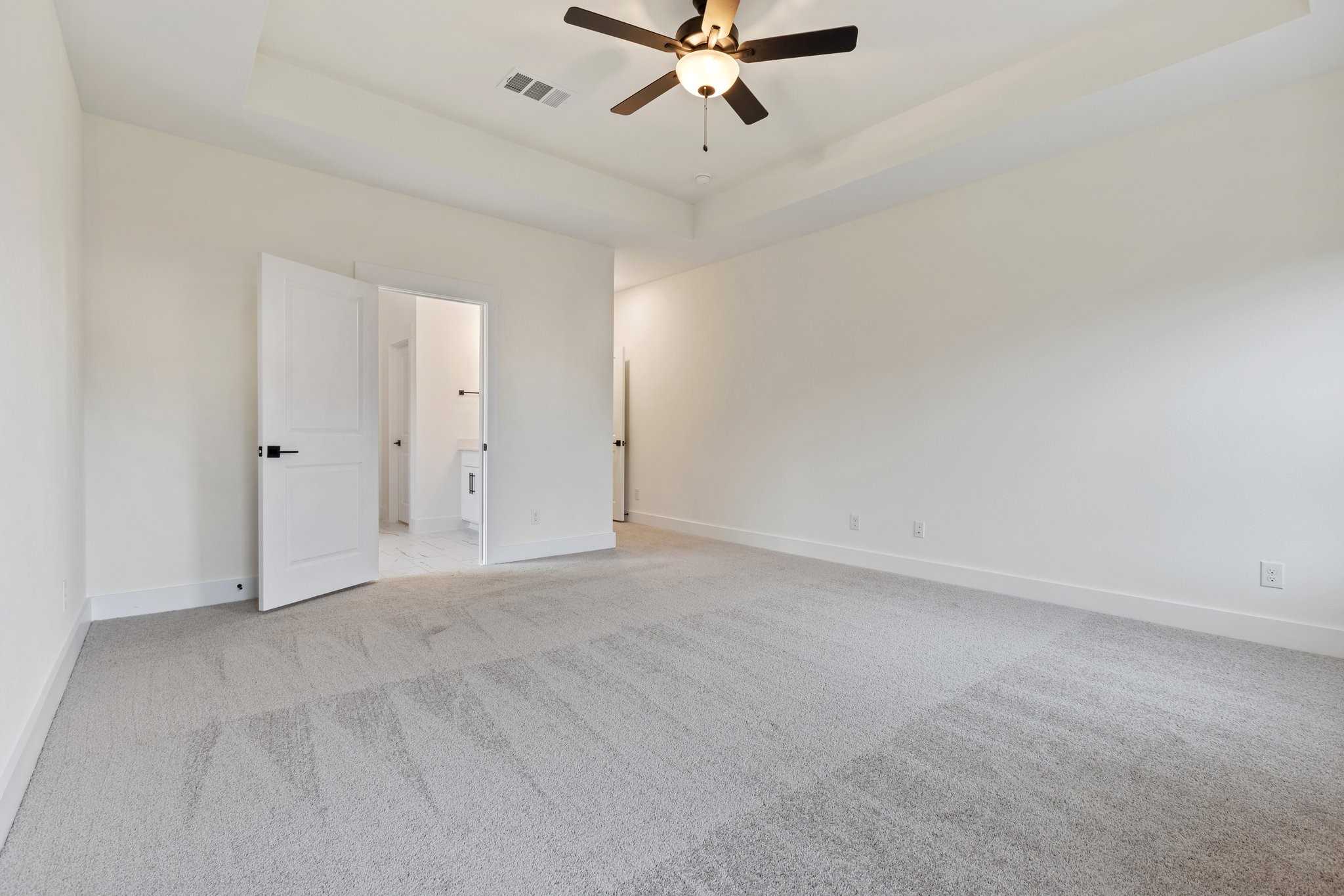 Spacious master bedroom with open doors to double vanity bathroom, tray ceiling fan, and beige carpet in Davidson Homes The Rockford C, Josephine, Texas
