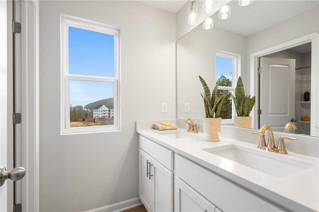 Modern double-sink vanity bathroom with gold fixtures, potted plant, and scenic window view in Davidson Homes The Wilmington B, Emerson, Georgia