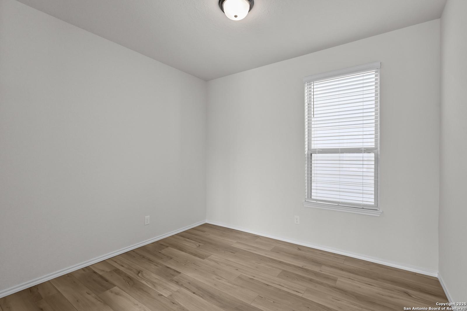 Bright secondary bedroom with white walls, blinds window, ceiling light, and hardwood floors in Davidson Homes Florence C, San Antonio