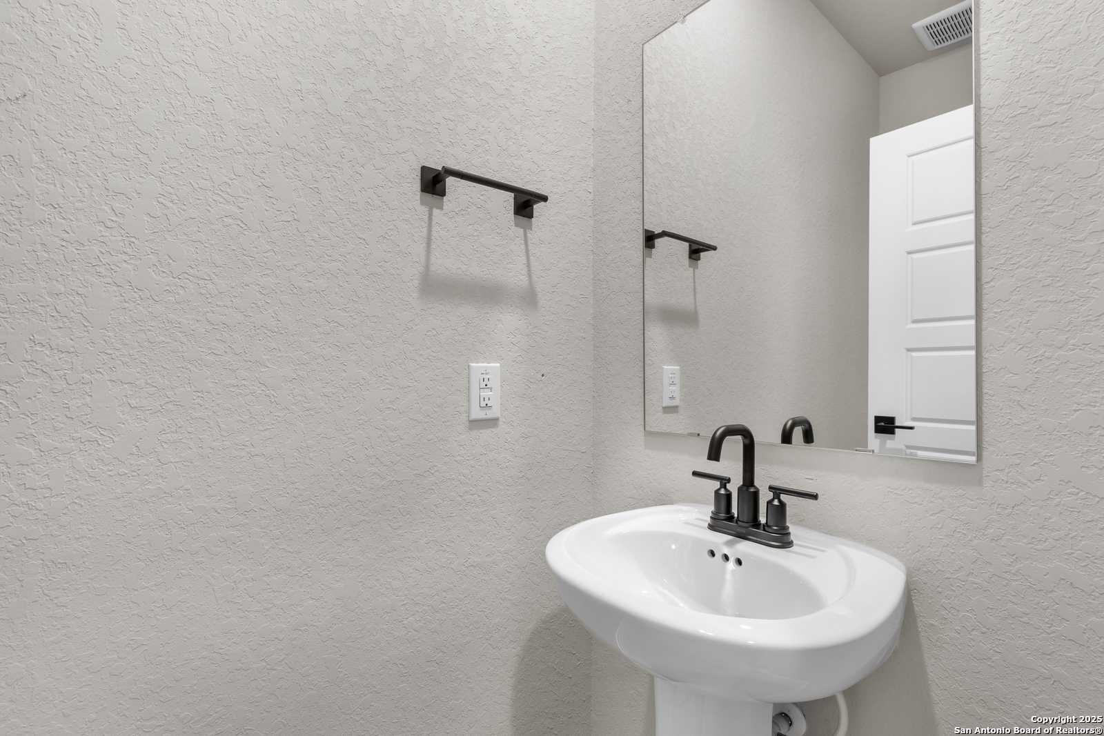 Elegant powder room with white pedestal sink, black faucet, and frameless mirror in Davidson Homes The Murray I, San Antonio