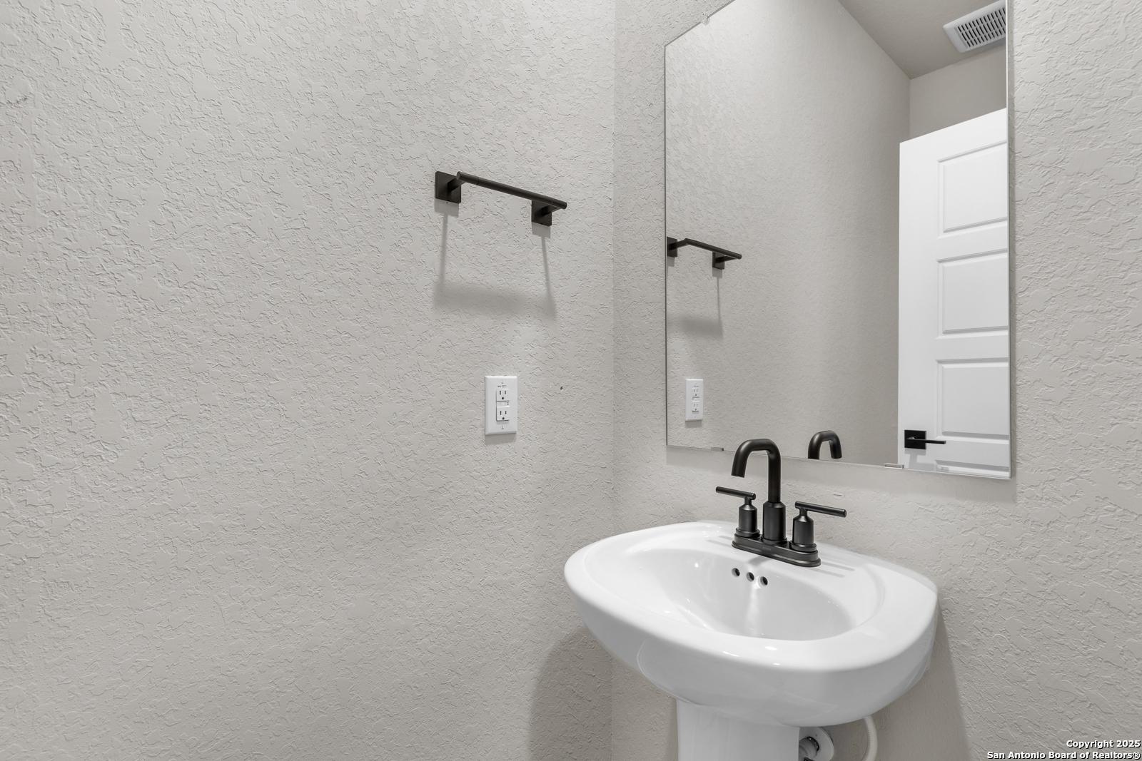 Elegant powder room with white pedestal sink, black faucet, and frameless mirror in Davidson Homes The Murray I, San Antonio
